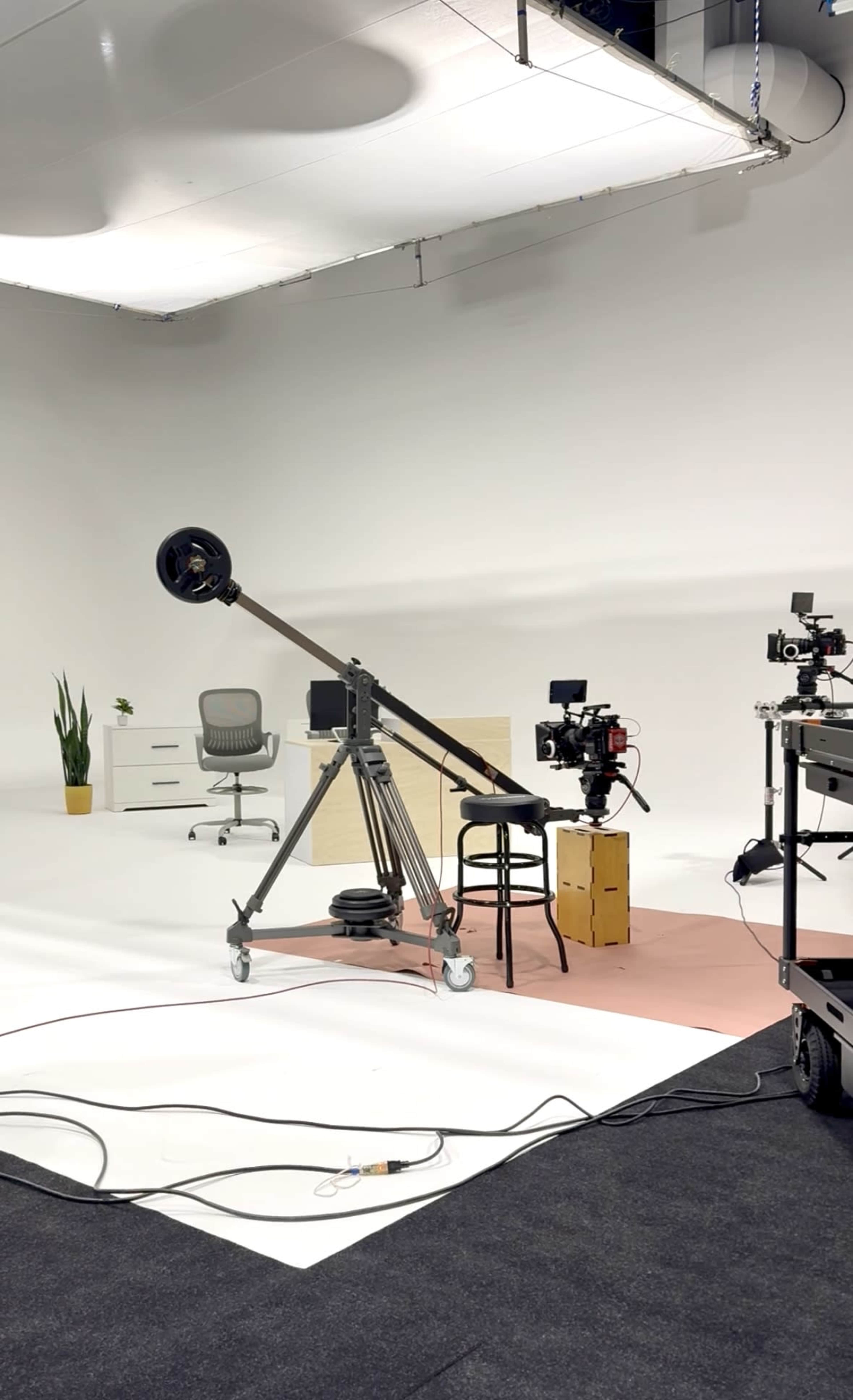 A film studio setup features a camera on a dolly track, a chair, a desk, and various filming equipment in a minimalistic white space.