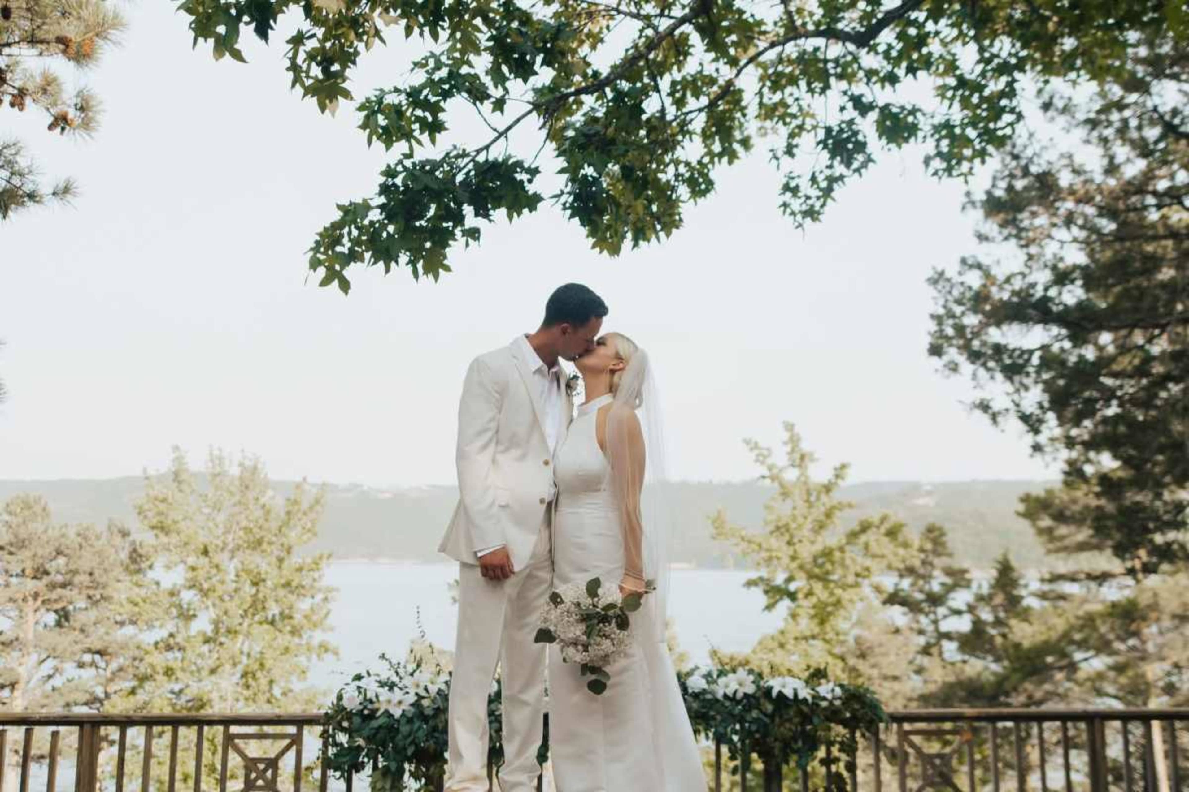 A bride in a white dress and a groom in a white suit share a kiss under a tree overlooking a serene body of water.