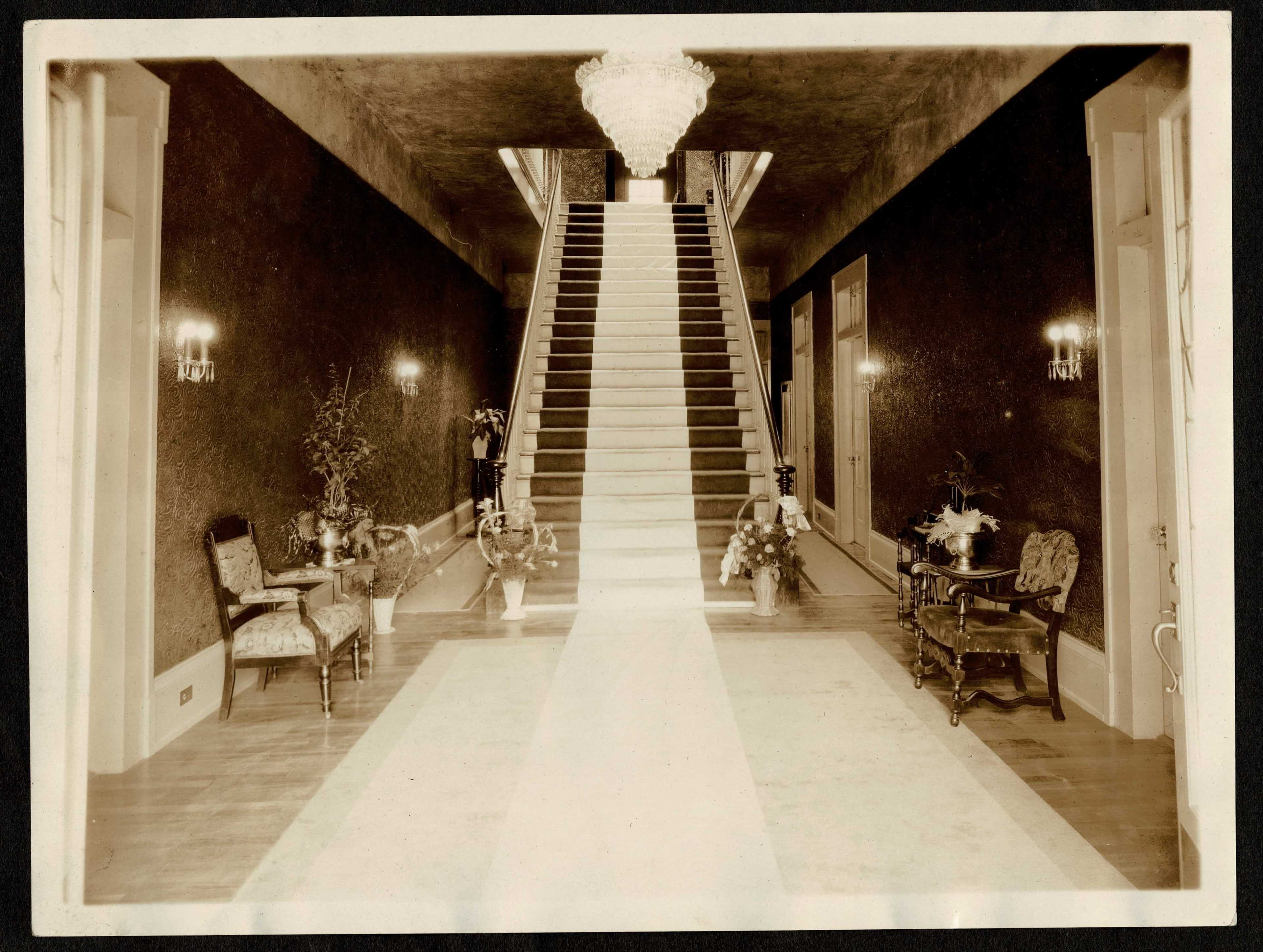 The image shows a grand staircase in a well-decorated hallway, flanked by ornate furniture and floral arrangements.