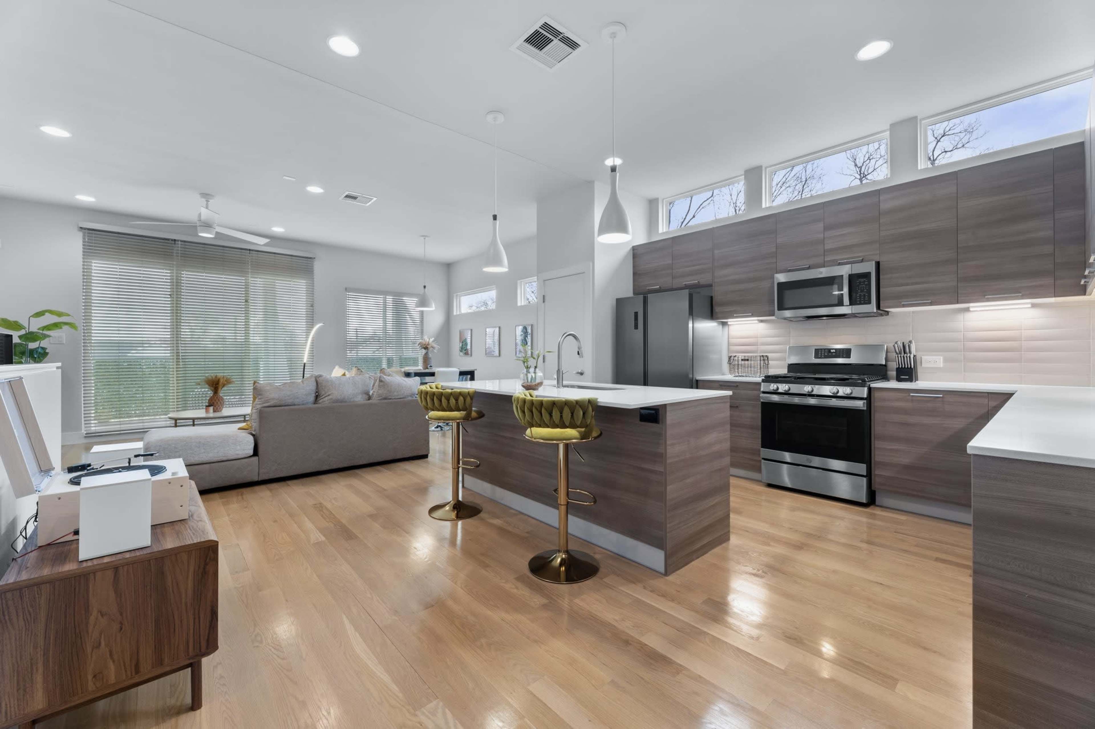 A modern kitchen with wooden cabinets, stainless steel appliances, and a countertop facing a living area with a couch and large windows.