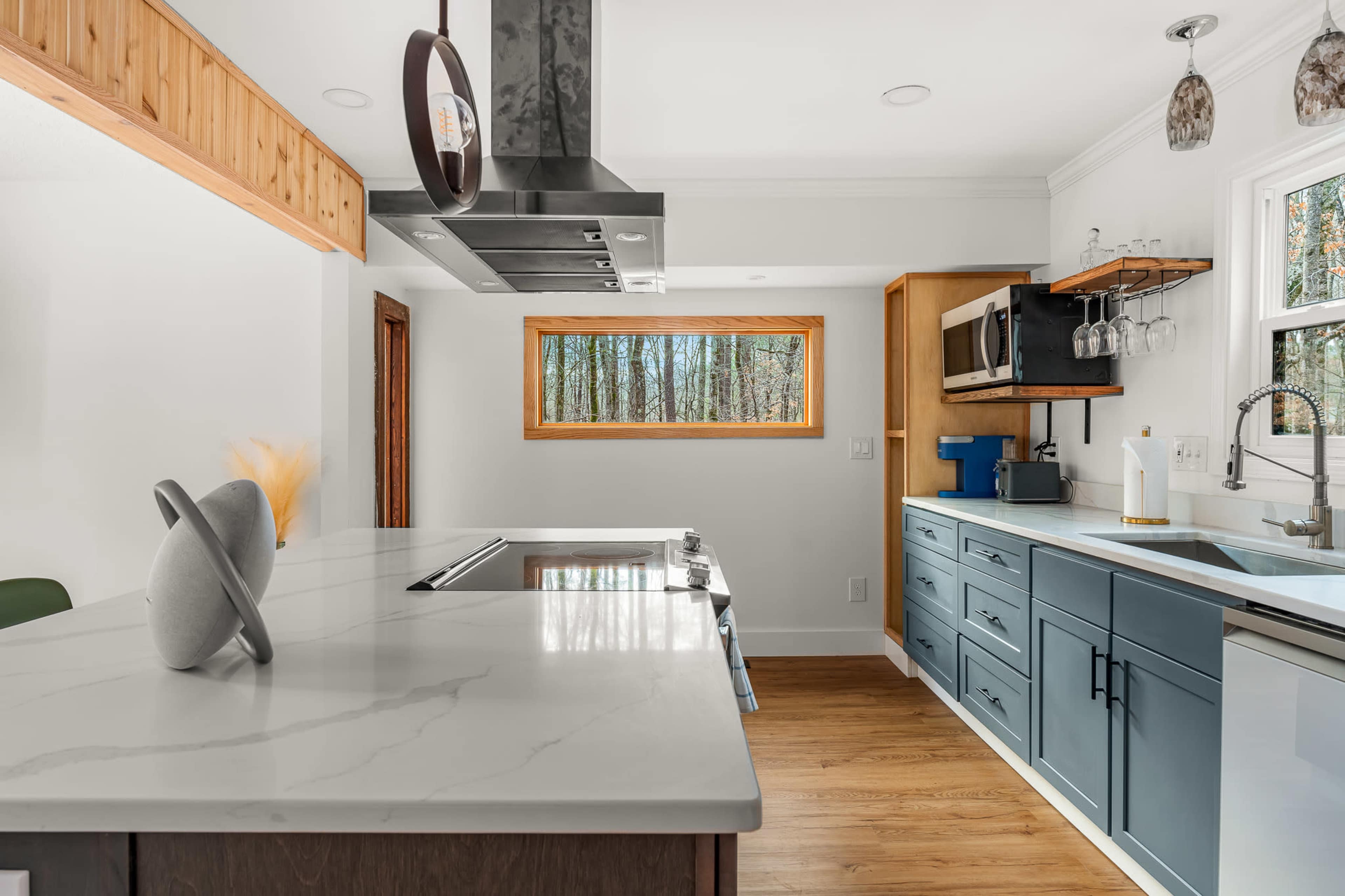 A modern kitchen features a central island with a stovetop, blue cabinets, and a large window showcasing a view of trees outside.