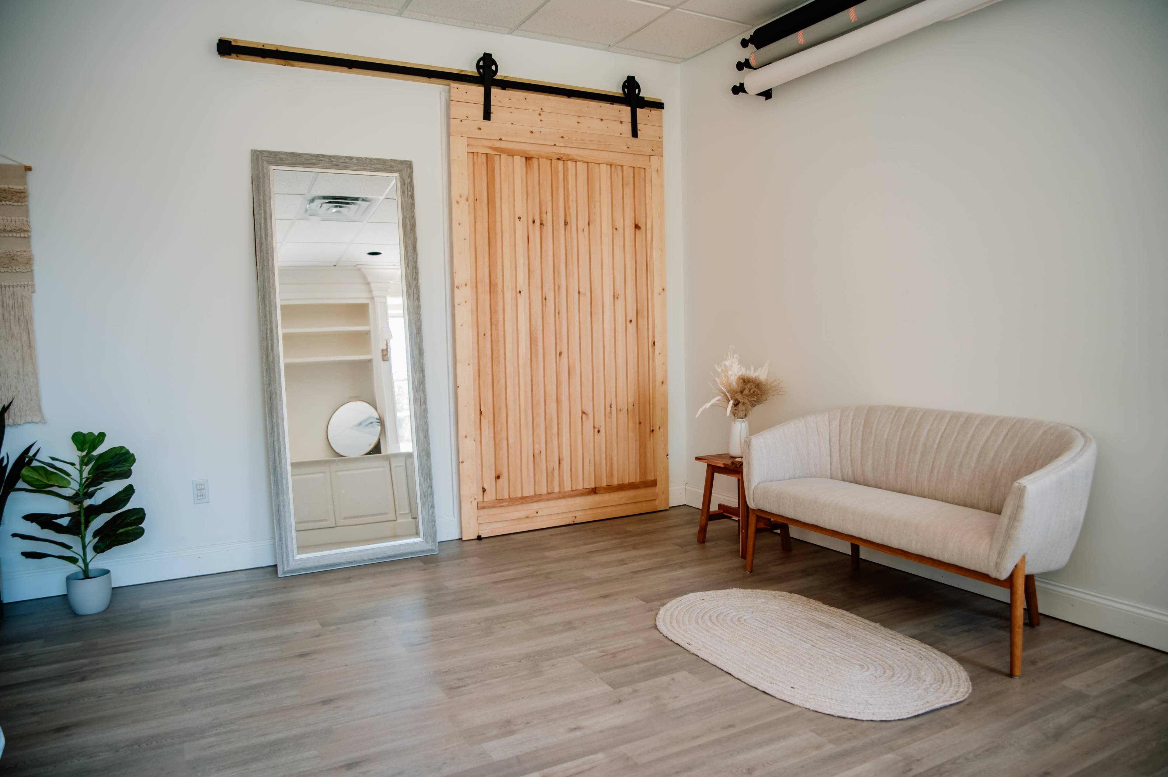 A minimalist interior space featuring a wooden sliding door, a full-length mirror, a light-colored couch, a small side table, and a round rug on a wooden floor.