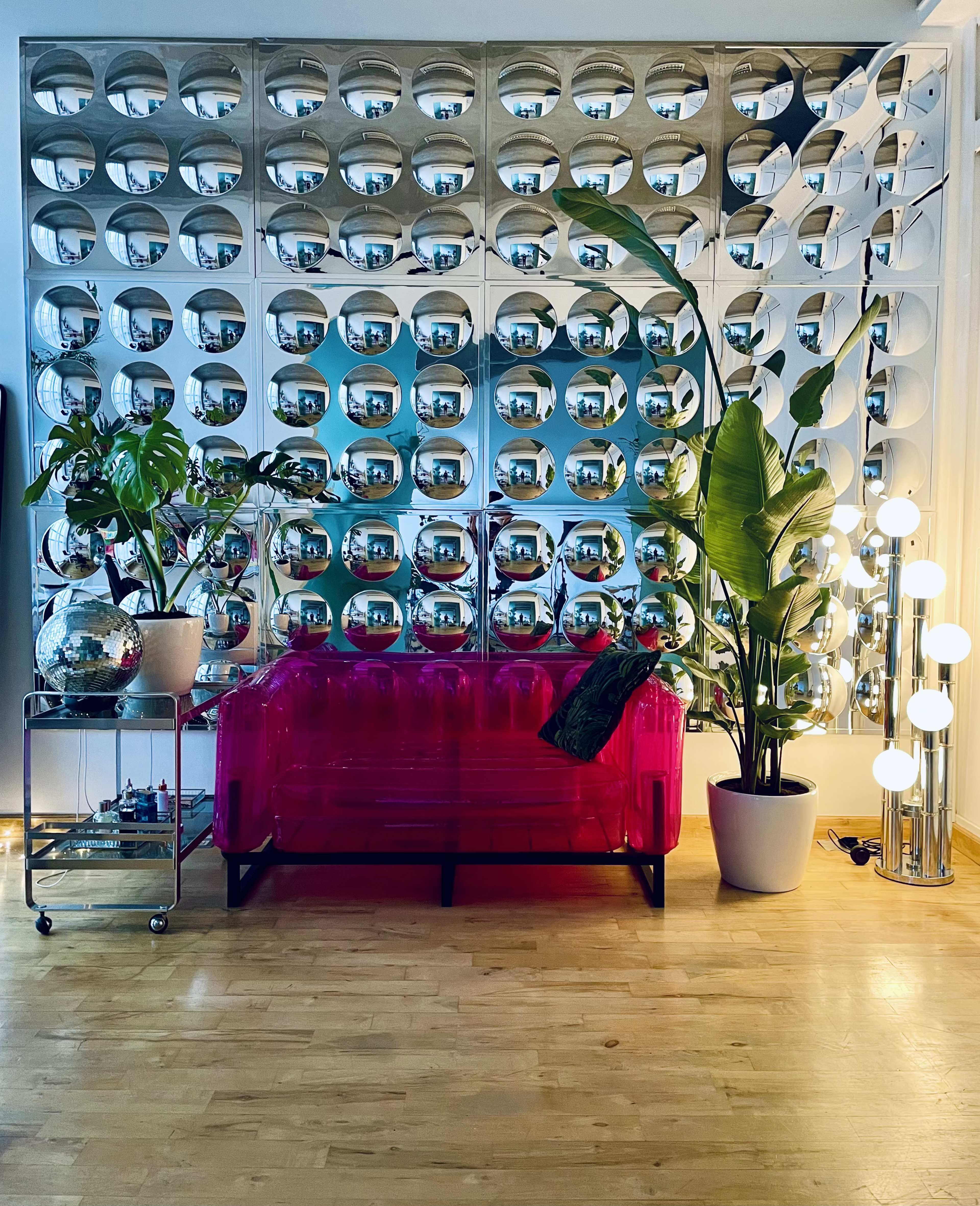The image features a pink transparent couch in front of a wall covered with circular mirrors, accompanied by potted plants and a lamp with multiple spherical bulbs.