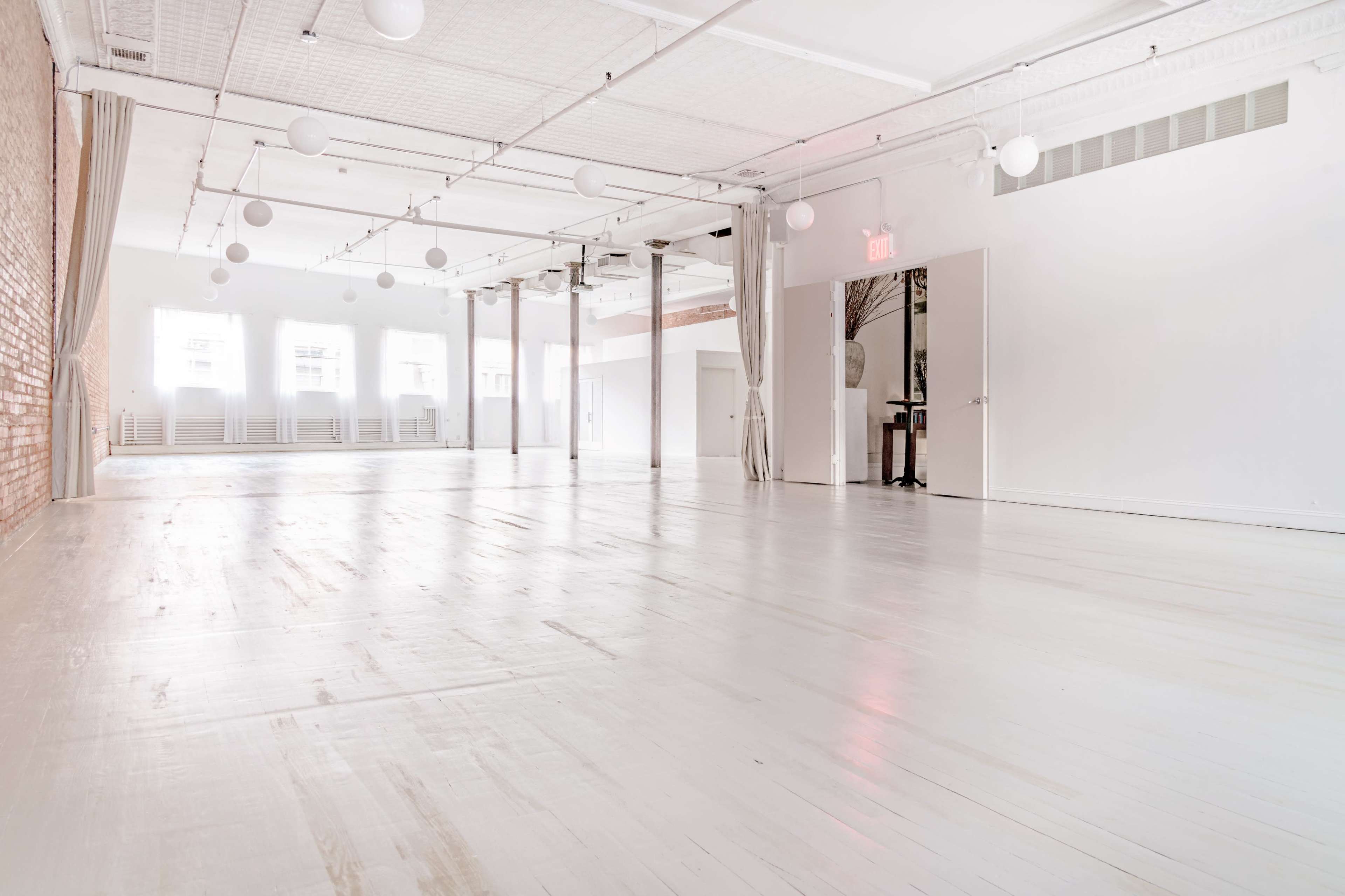 The image shows a spacious, well-lit interior room with wooden floors, white walls, and hanging spherical light fixtures.