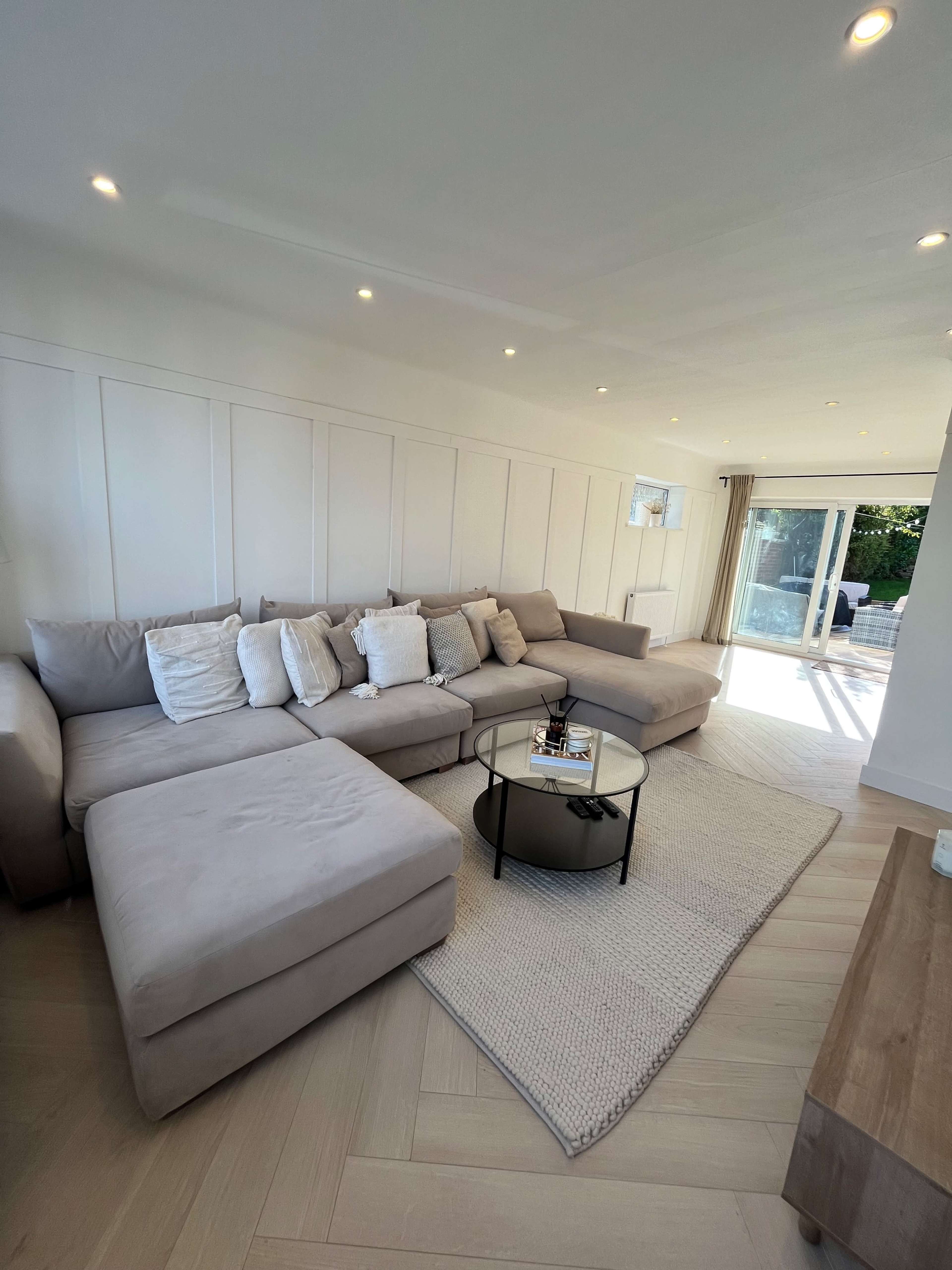 A modern living room featuring a large beige sectional sofa, a circular glass coffee table, and natural light streaming in from a nearby sliding door.