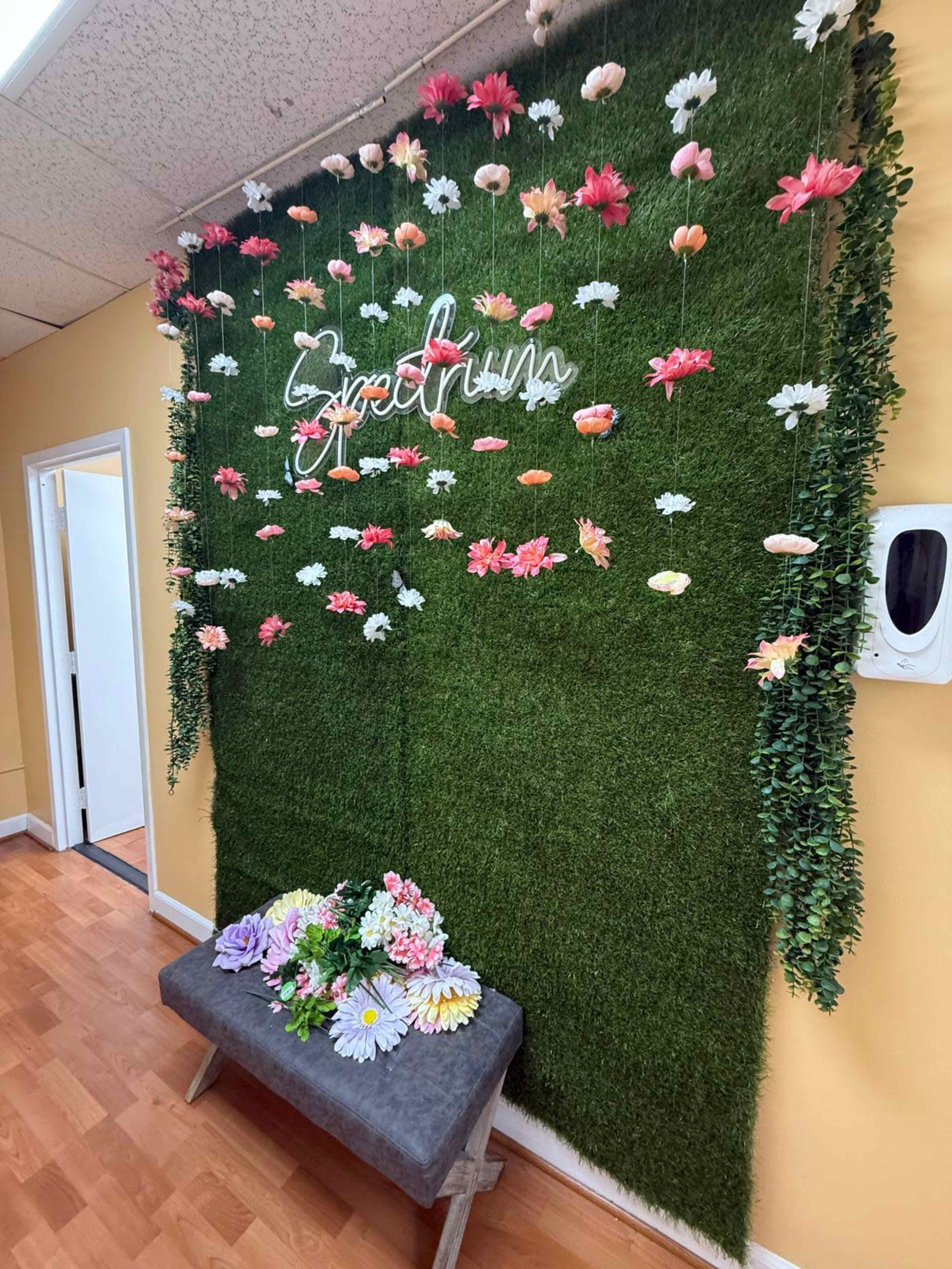 A vibrant wall decorated with artificial flowers and greenery, featuring a neon sign that reads "Sprout," alongside a bench adorned with fresh-looking artificial flowers.