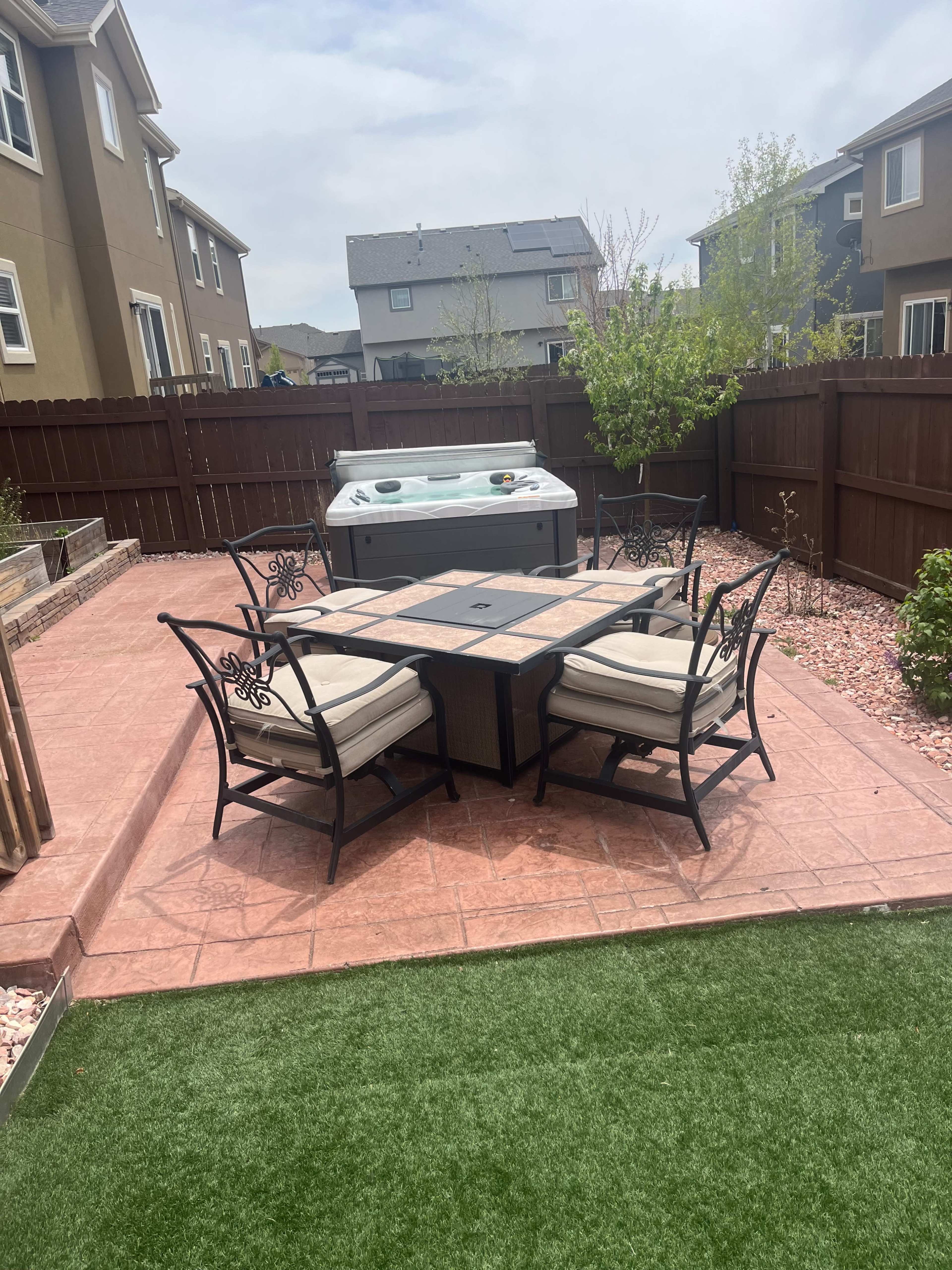 A backyard patio features a dining table surrounded by chairs and a hot tub, with a landscaped area of grass and stone.