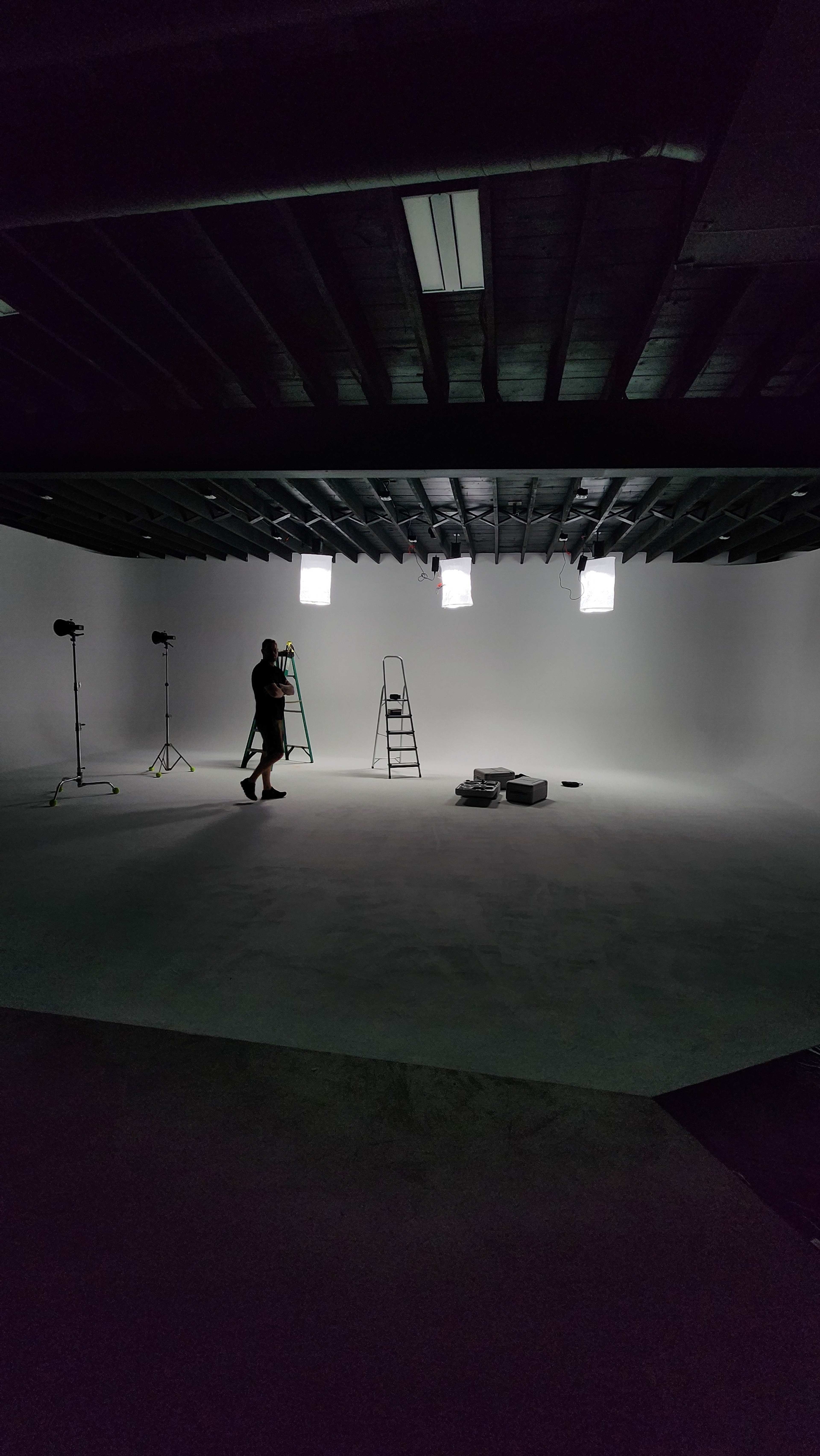 A person stands on a ladder in a dimly lit studio space with bright lights mounted above.