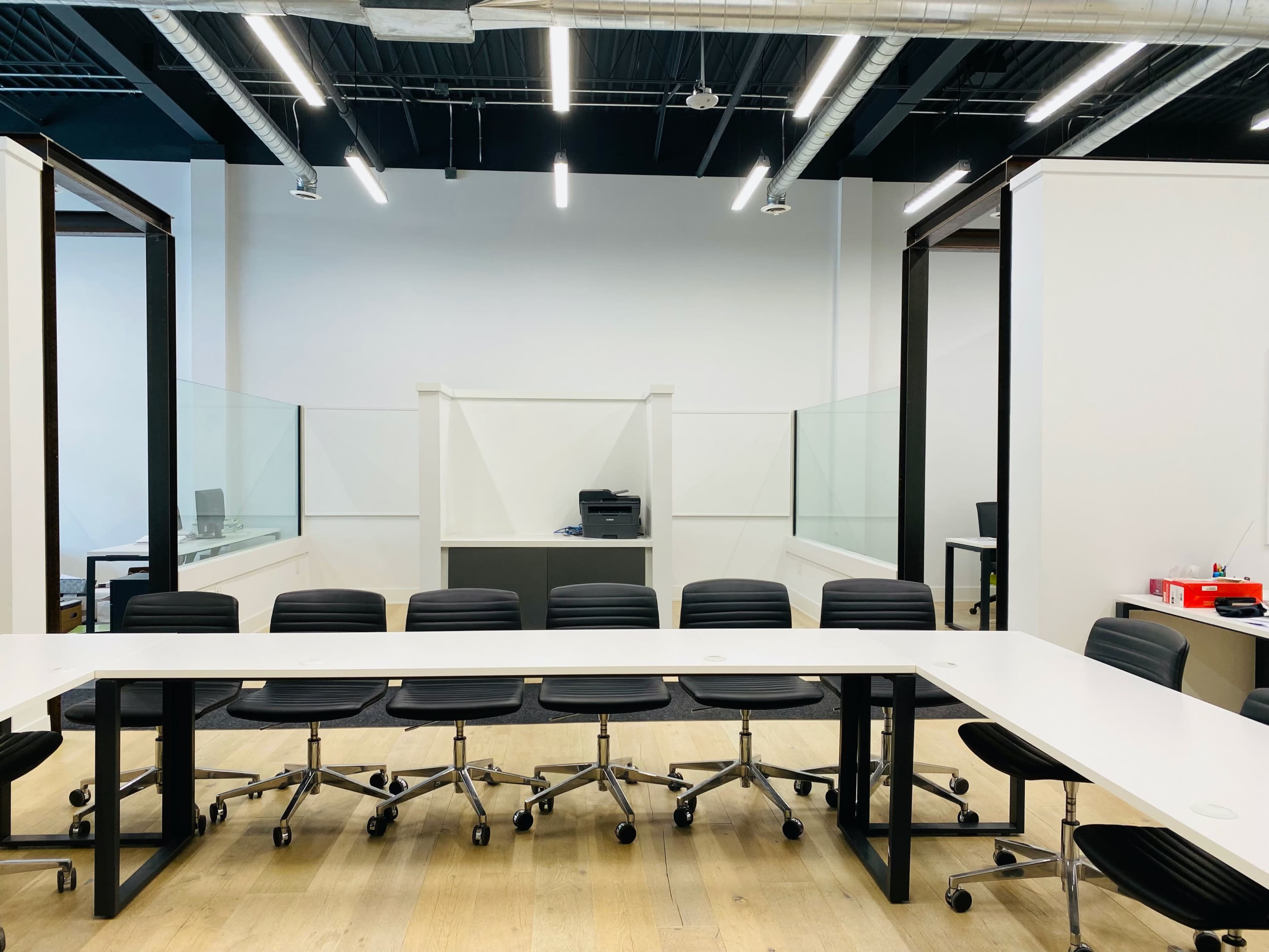 A spacious conference room features a long white table with black rolling chairs and a backdrop of glass partitions and office equipment.