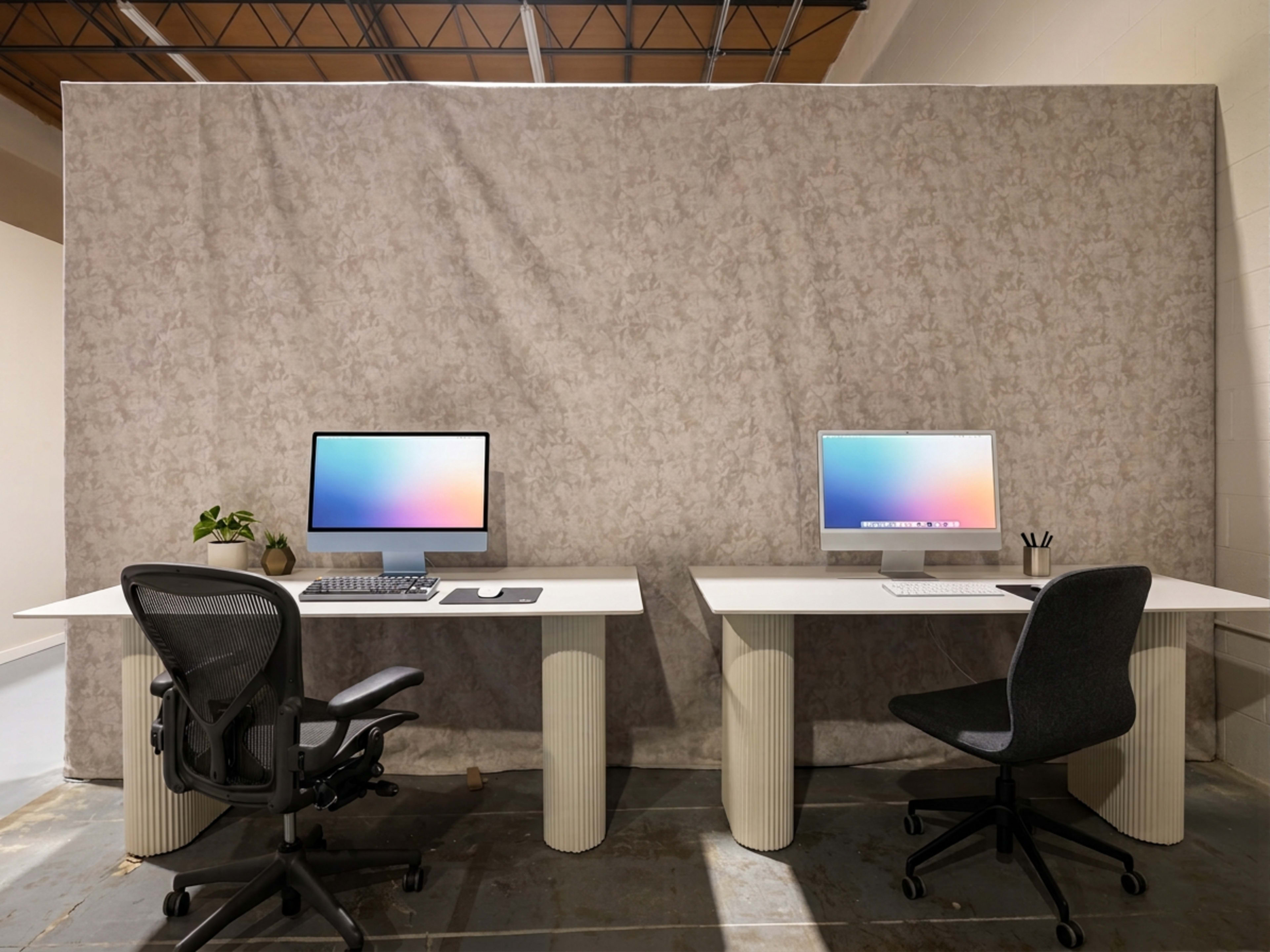 Two modern desks with computers are positioned side by side in front of a textured gray wall, each equipped with office chairs and minimal decorative elements.