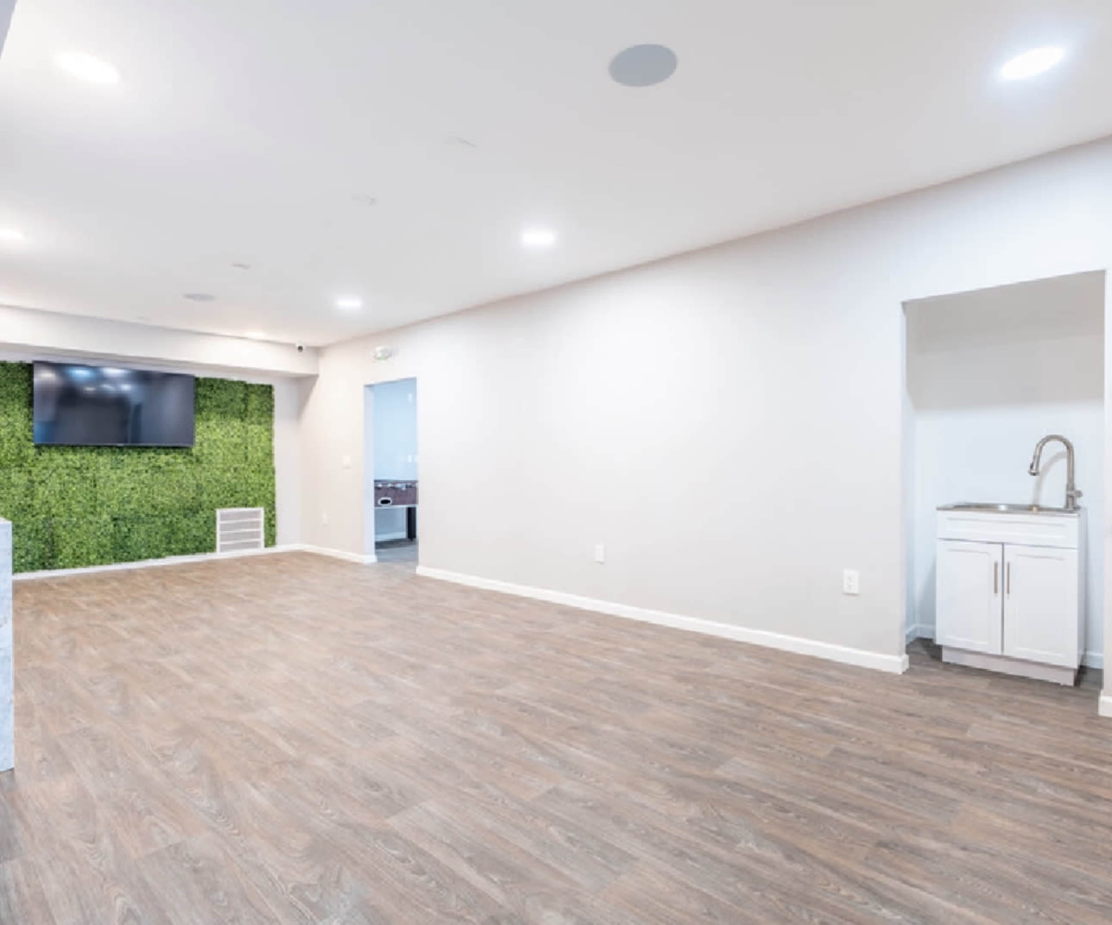 A spacious, empty room with wooden flooring, a green wall feature, and a small kitchenette in the corner.
