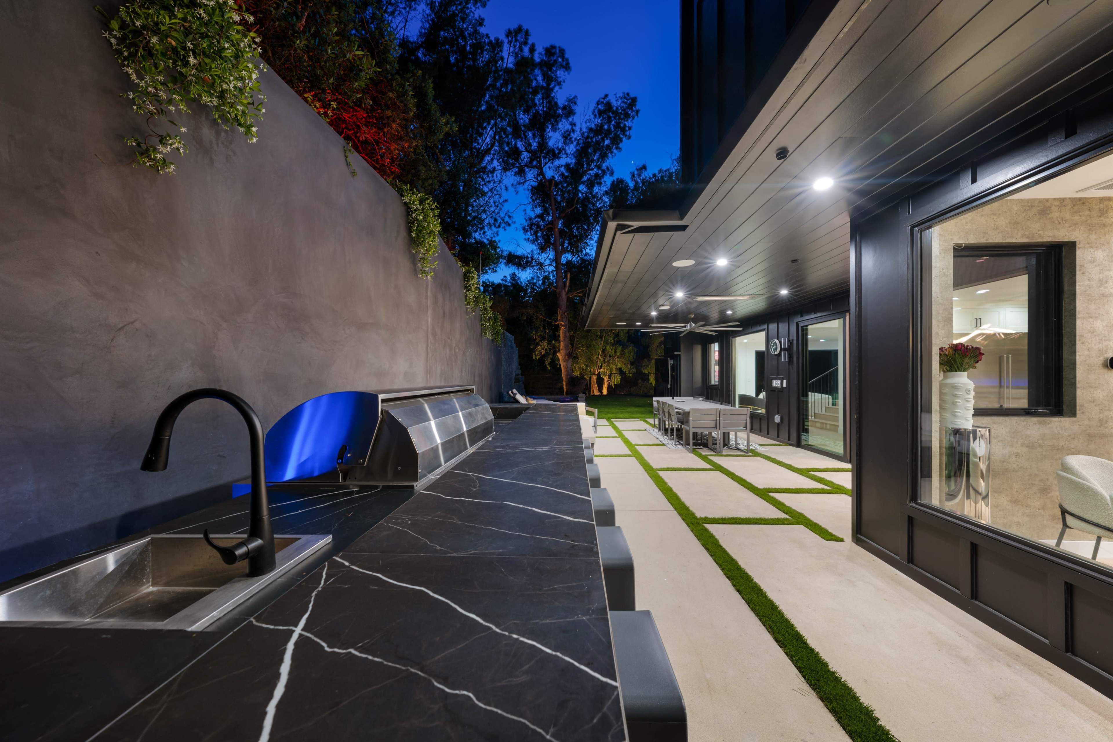 The image shows a modern outdoor kitchen area featuring a stone countertop, a built-in grill, and a spacious patio with seating and landscaping under evening lighting.