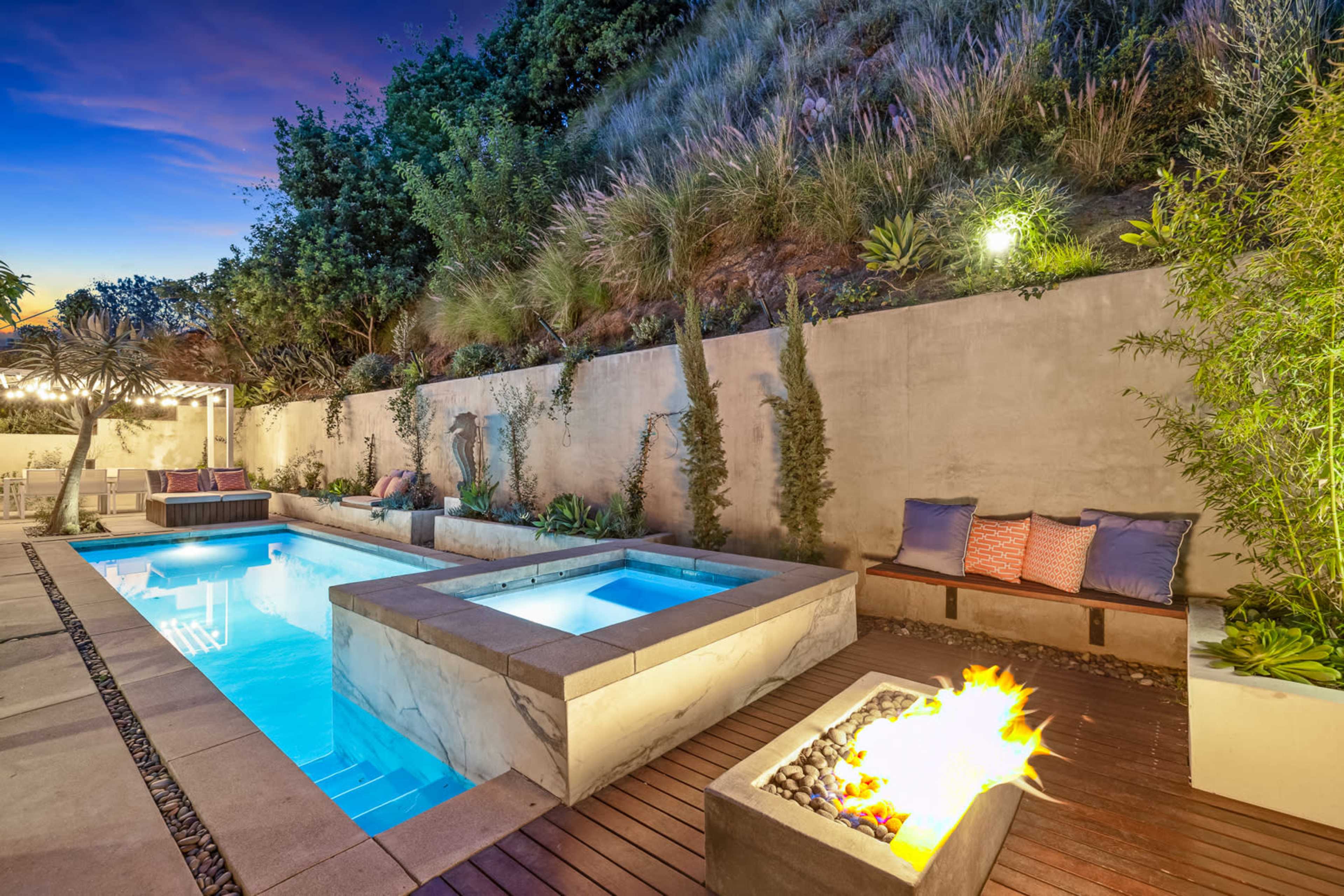 The image shows a modern outdoor space featuring a rectangular swimming pool, a spa, and a fire pit surrounded by landscaped greenery and seating areas.
