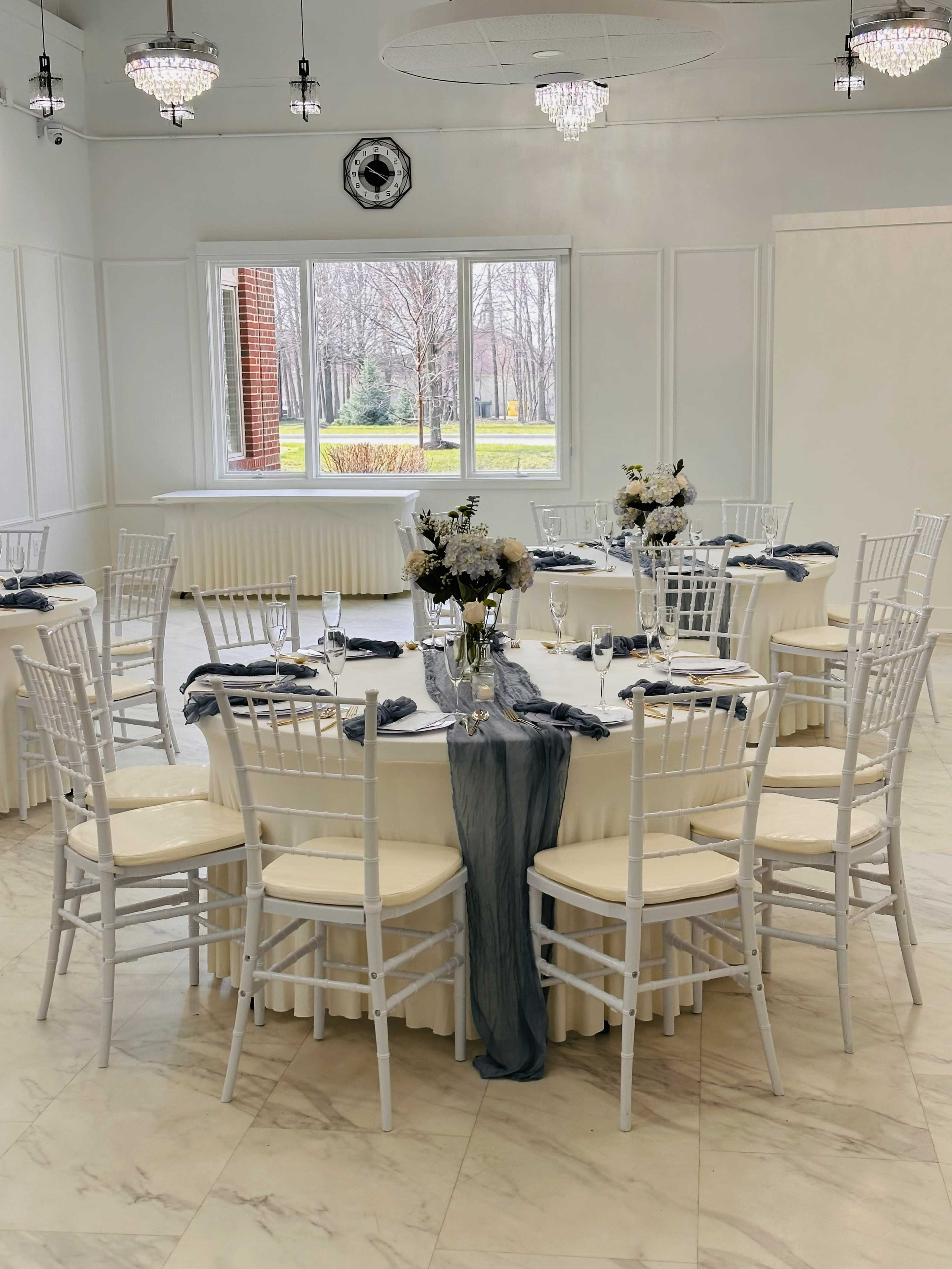 The image shows a decorated dining area with white tables set for a formal event, featuring floral centerpieces and elegant tableware.