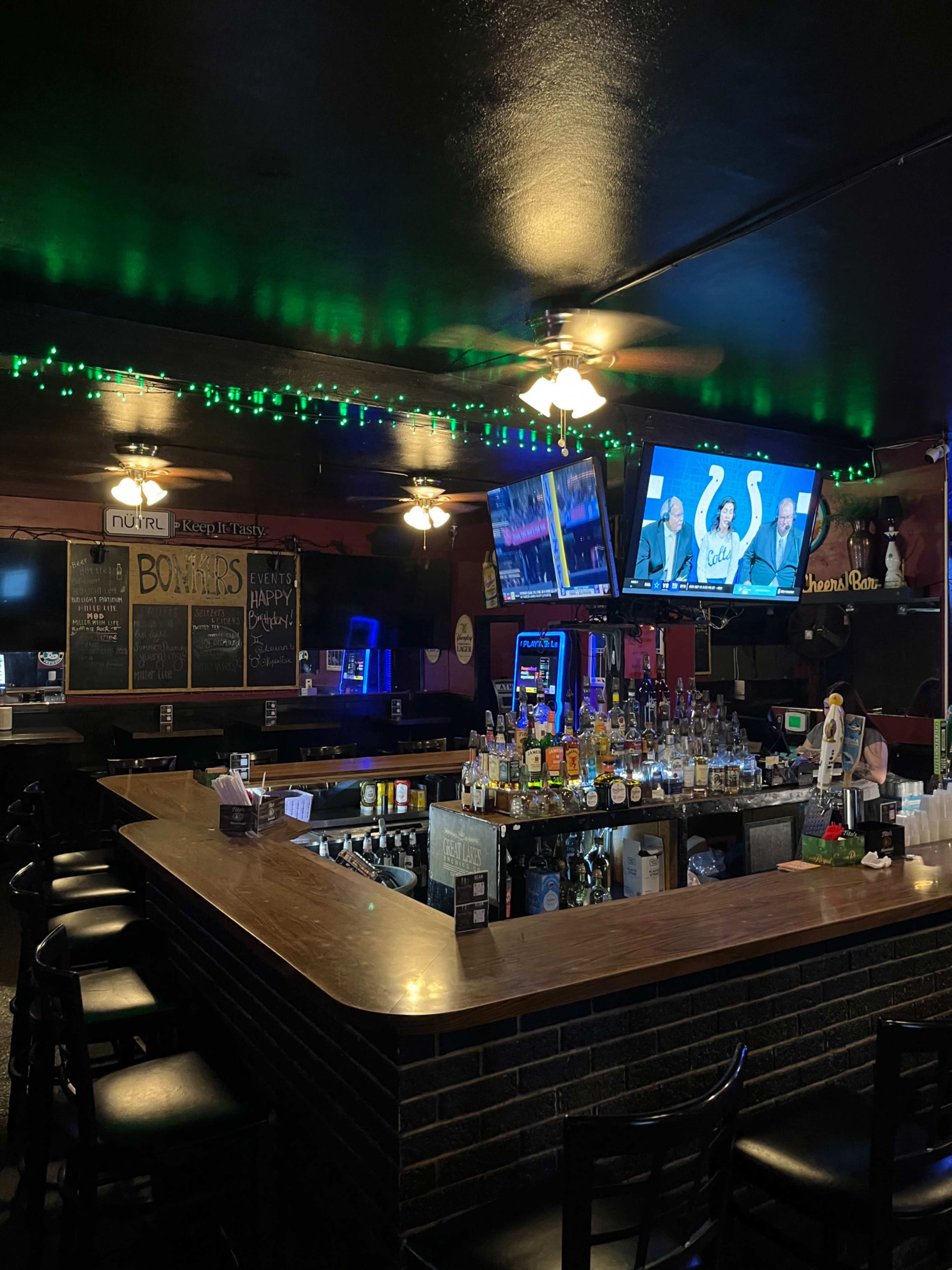The image shows the interior of a bar with a wooden countertop, several bar stools, and two televisions displaying content, illuminated by green lights along the ceiling.