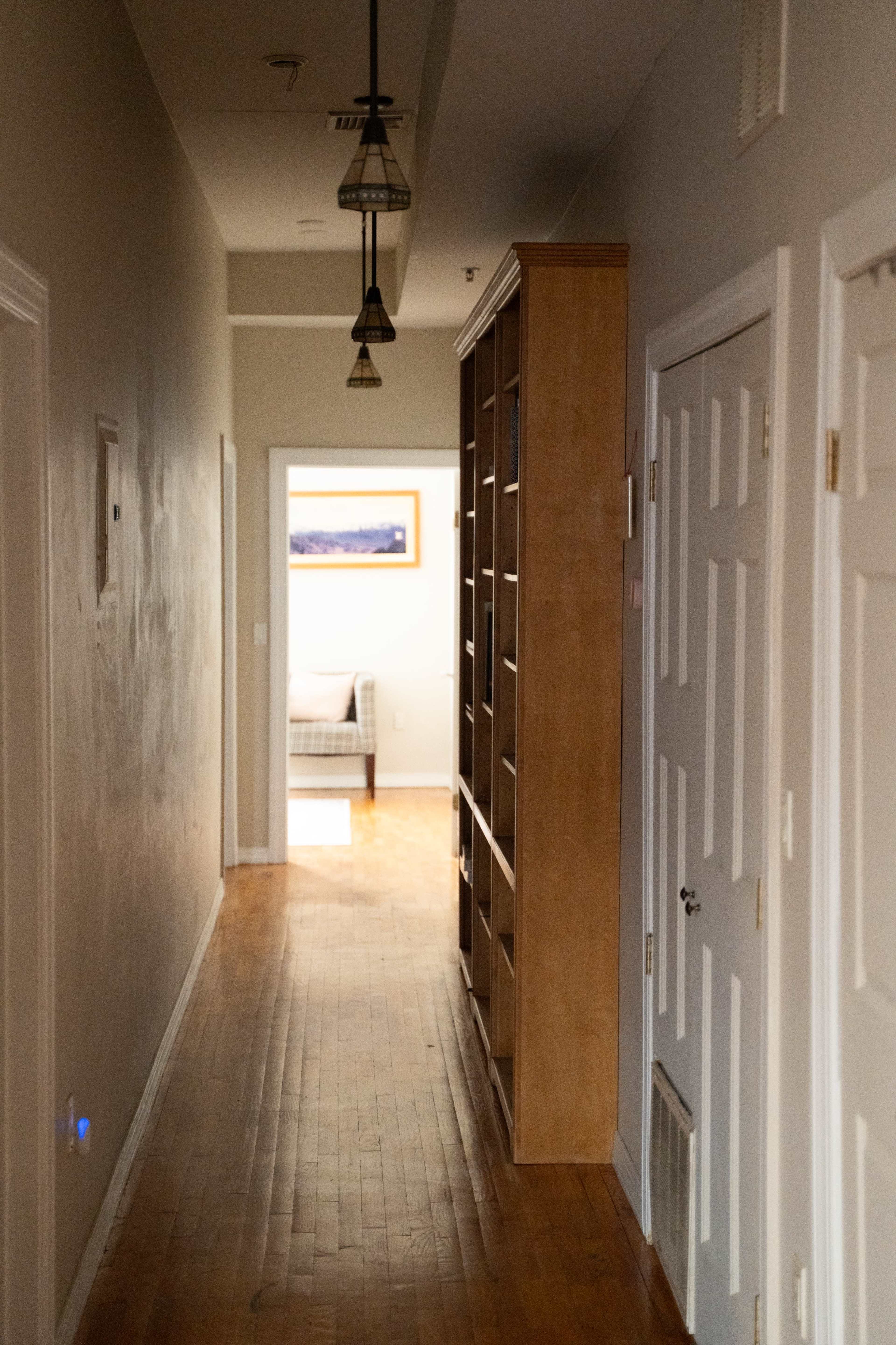 The image shows a narrow hallway with wooden flooring, a bookshelf on the left, and a doorway at the end leading to a room with furniture.