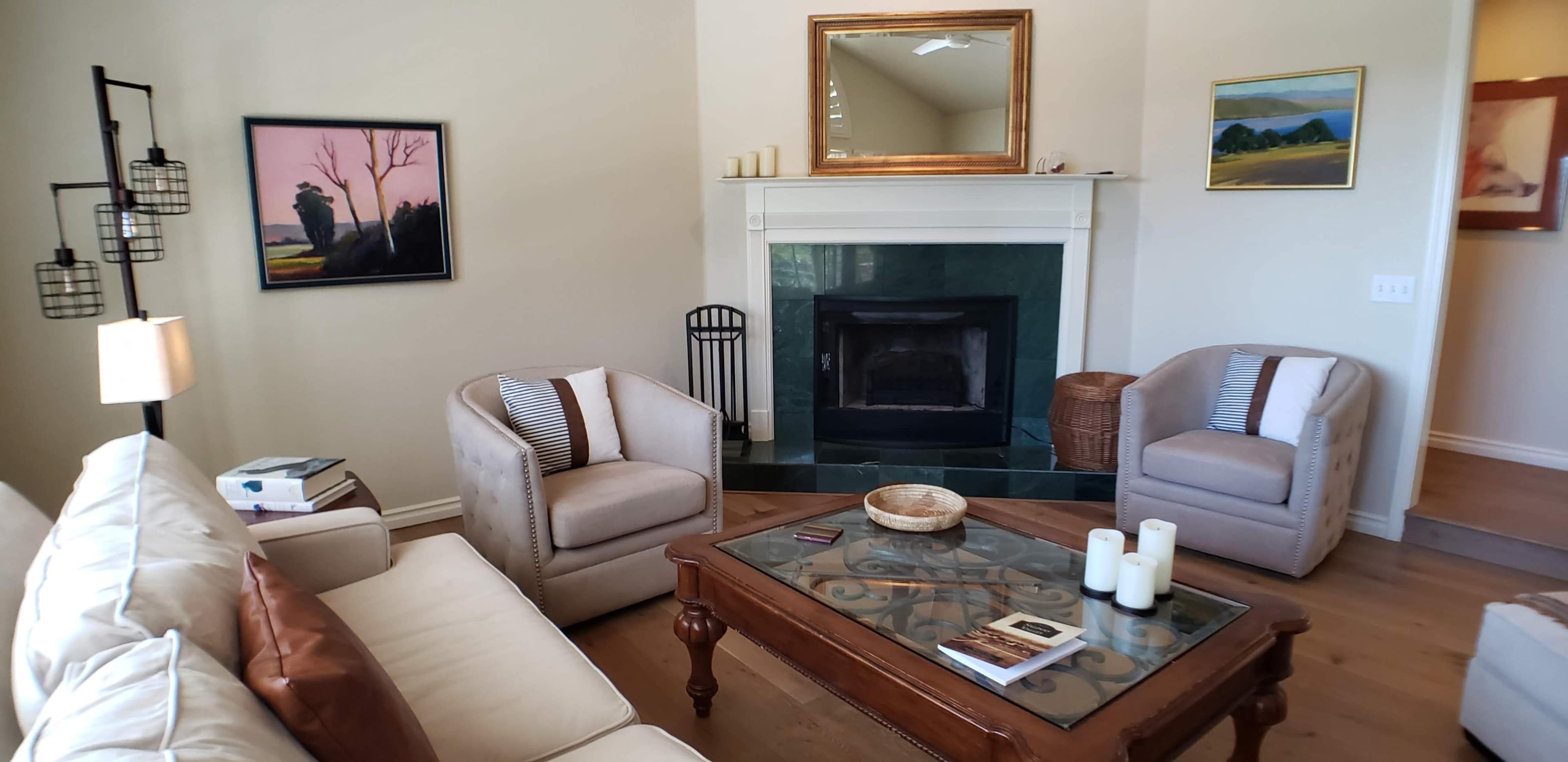The living room features a cream-colored sofa, two light gray armchairs, a wooden coffee table, and a fireplace with a green marble surround, set against neutral walls and decorated with artwork.