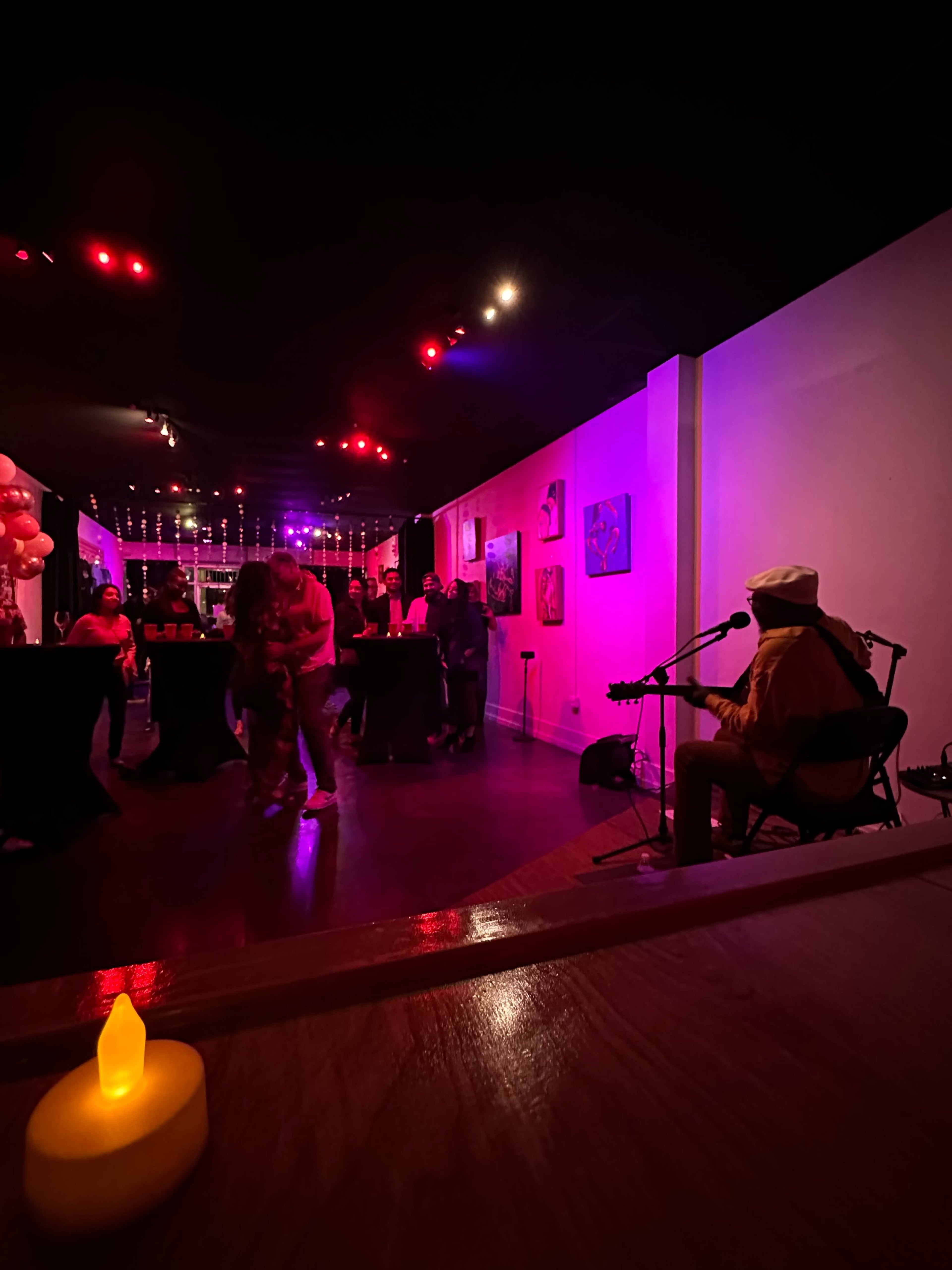 A musician plays guitar in a dimly lit venue with guests dancing and socializing around tables decorated with balloons.