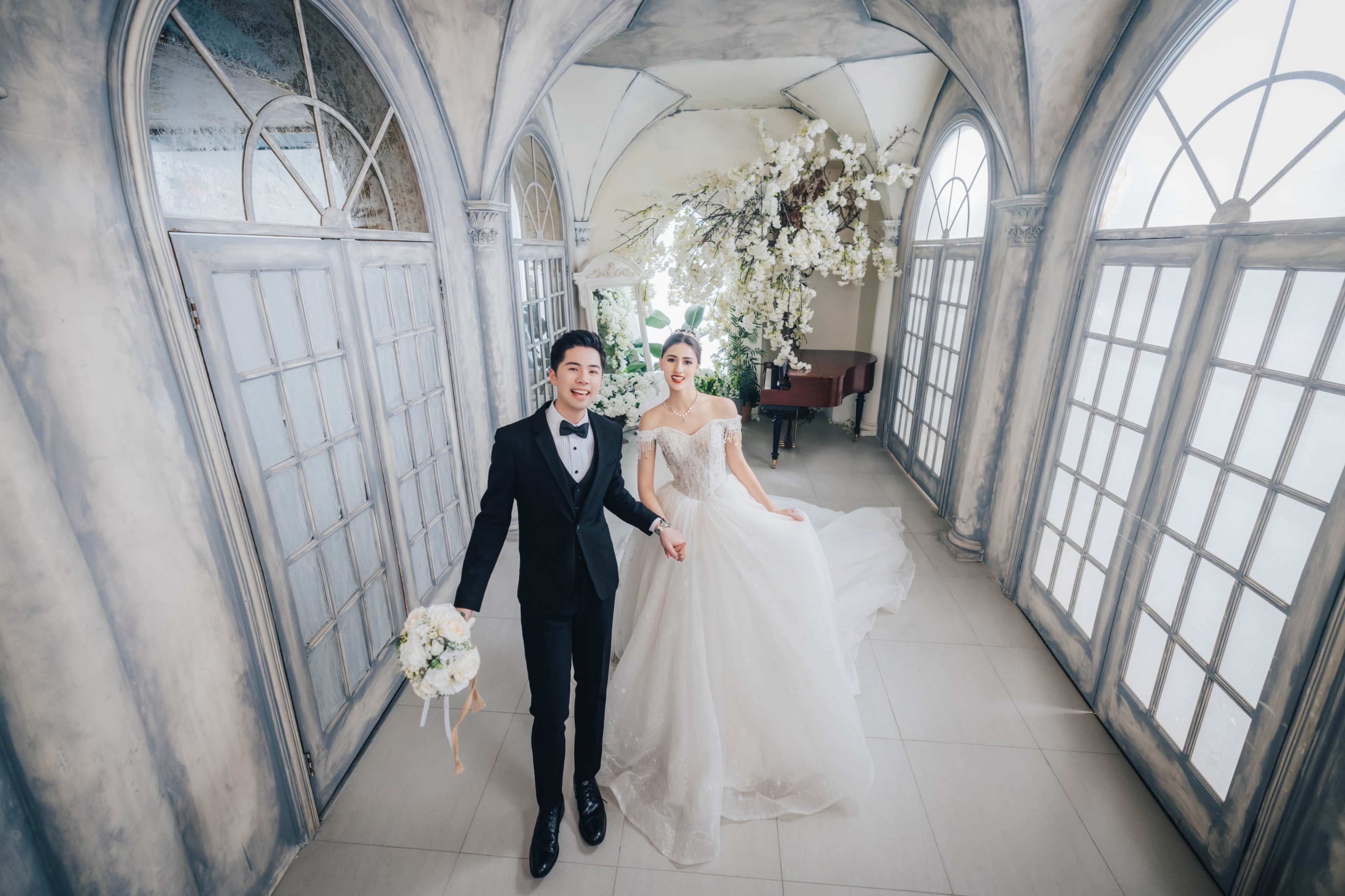 A bride and groom hold hands in a grand hall adorned with large arched windows and floral decorations.