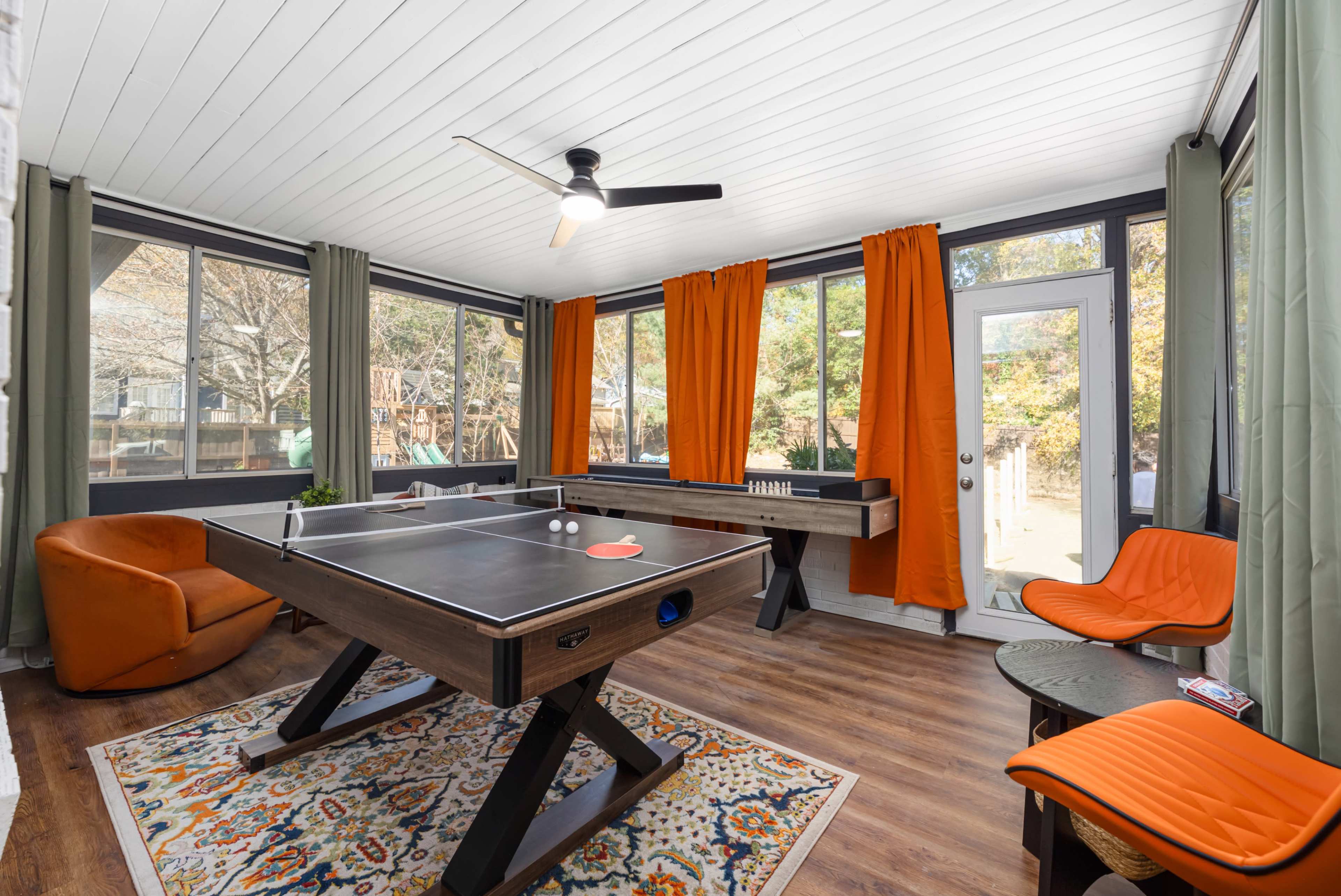 The image shows a well-lit sunroom featuring a ping pong table, an orange accent chair, a decorative rug, and large windows with green curtains.
