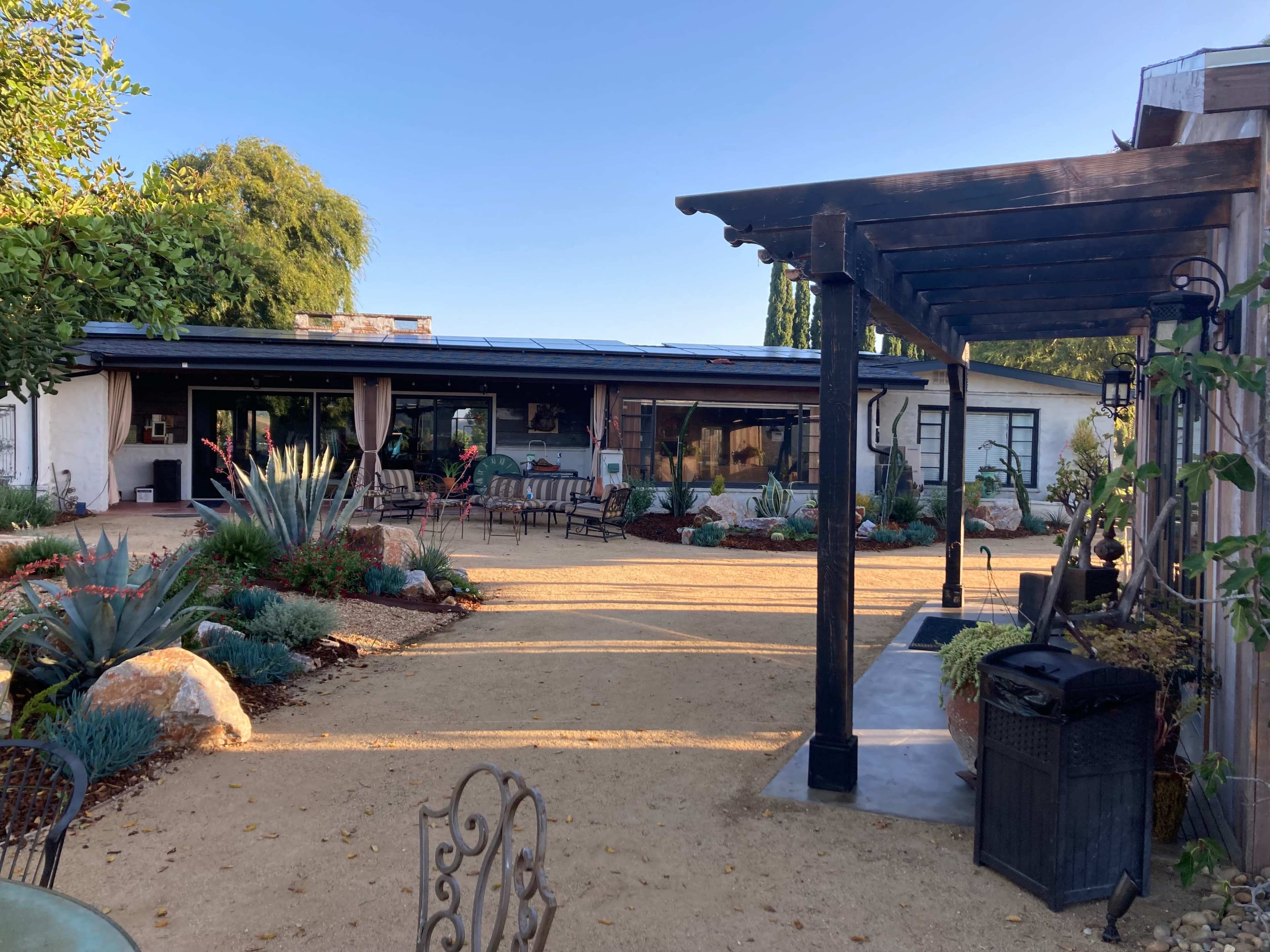 The image shows a landscaped courtyard with a gravel path leading to a modern building, framed by greenery and outdoor seating.