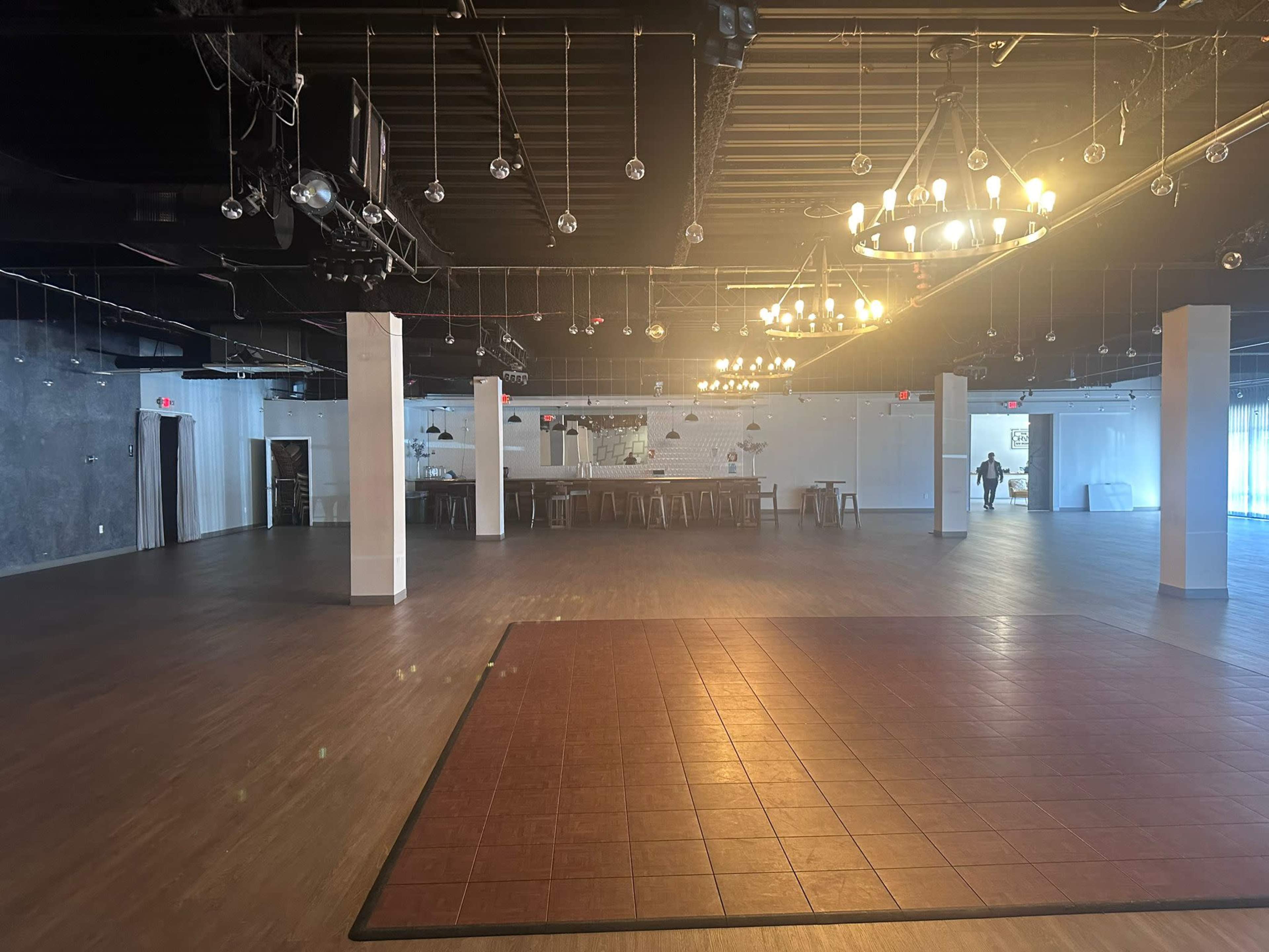 The image shows a spacious, empty event venue with wooden flooring, a large table in the center, and decorative lighting fixtures hanging from the ceiling.