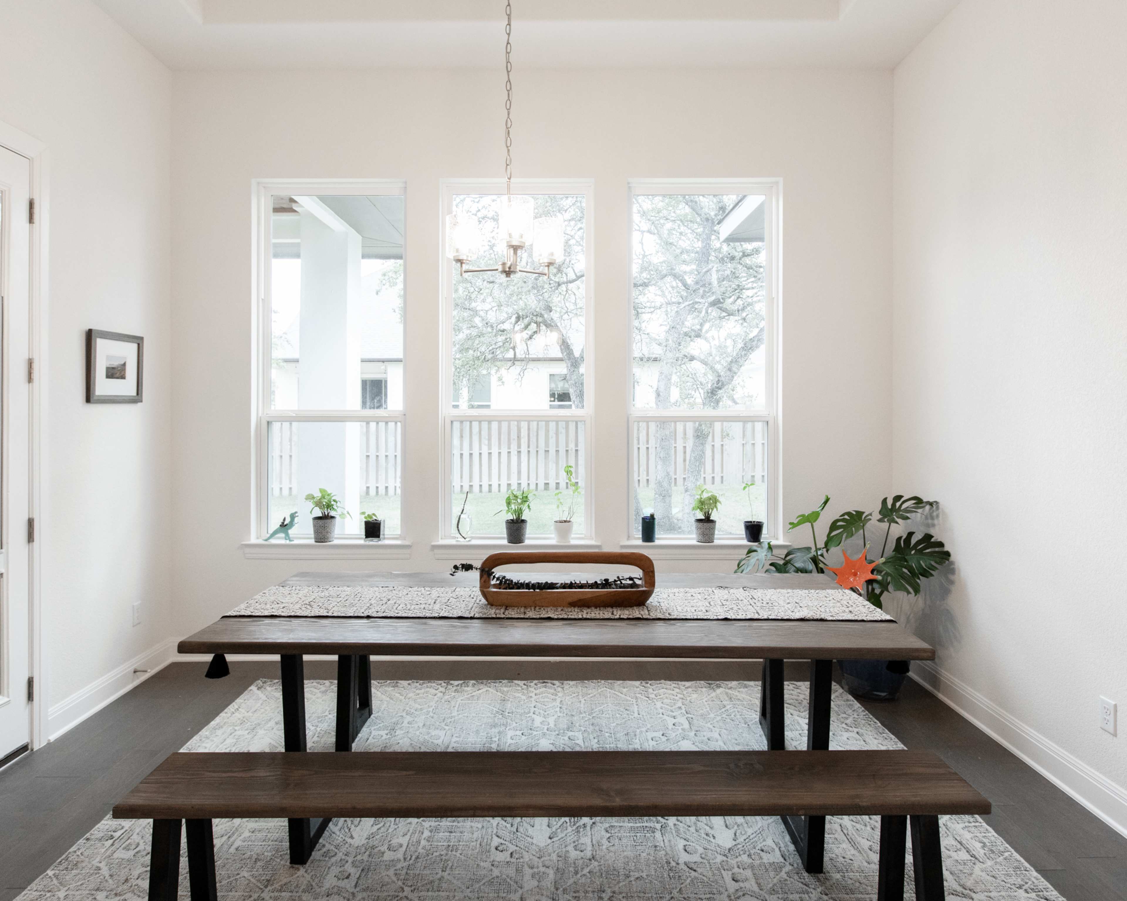A long wooden table with benches sits in a well-lit dining area, surrounded by windows with potted plants on the sills.