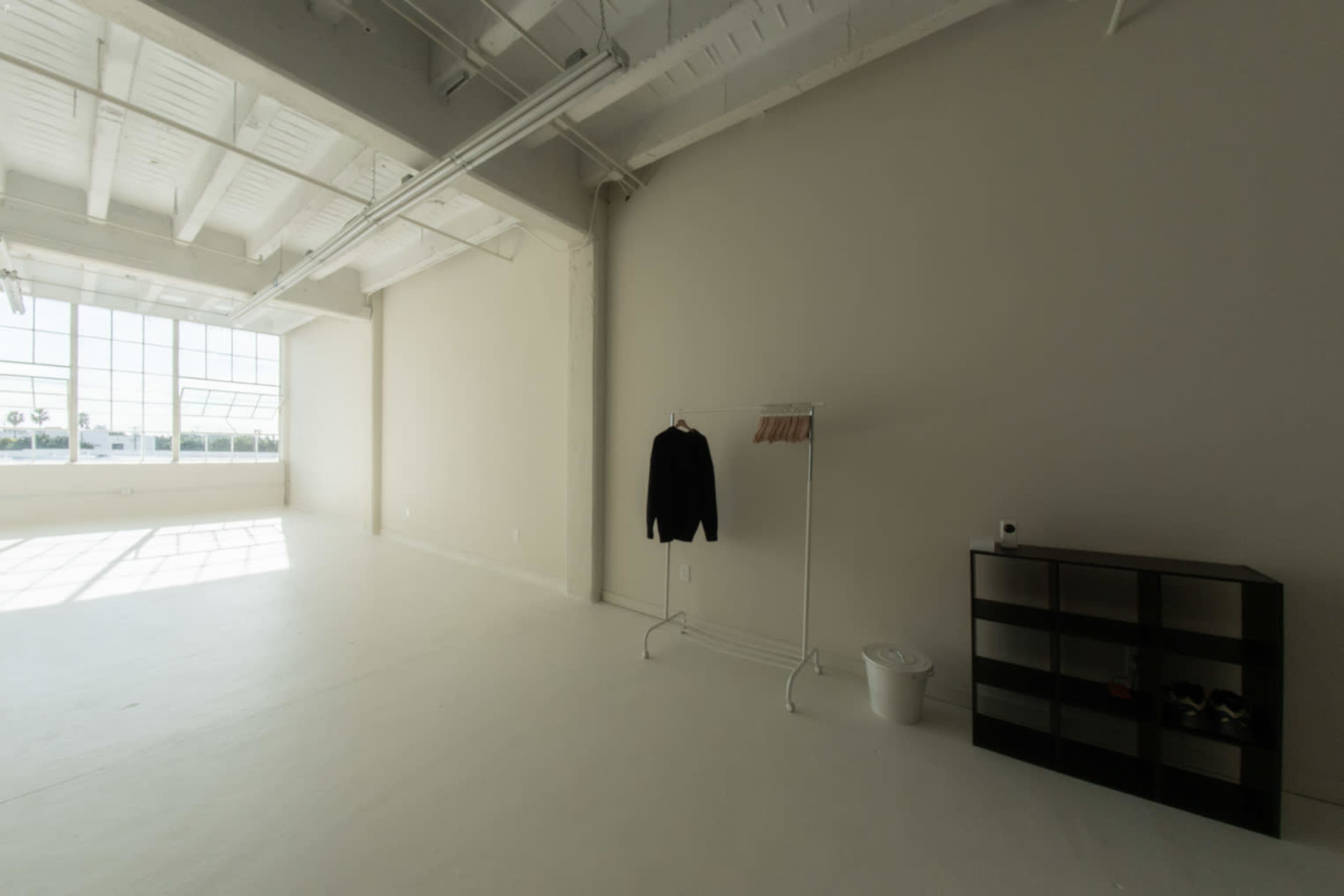 A spacious, empty room with white walls and a large window, featuring a clothing rack with a black garment, a white trash bin, and a black shelving unit.