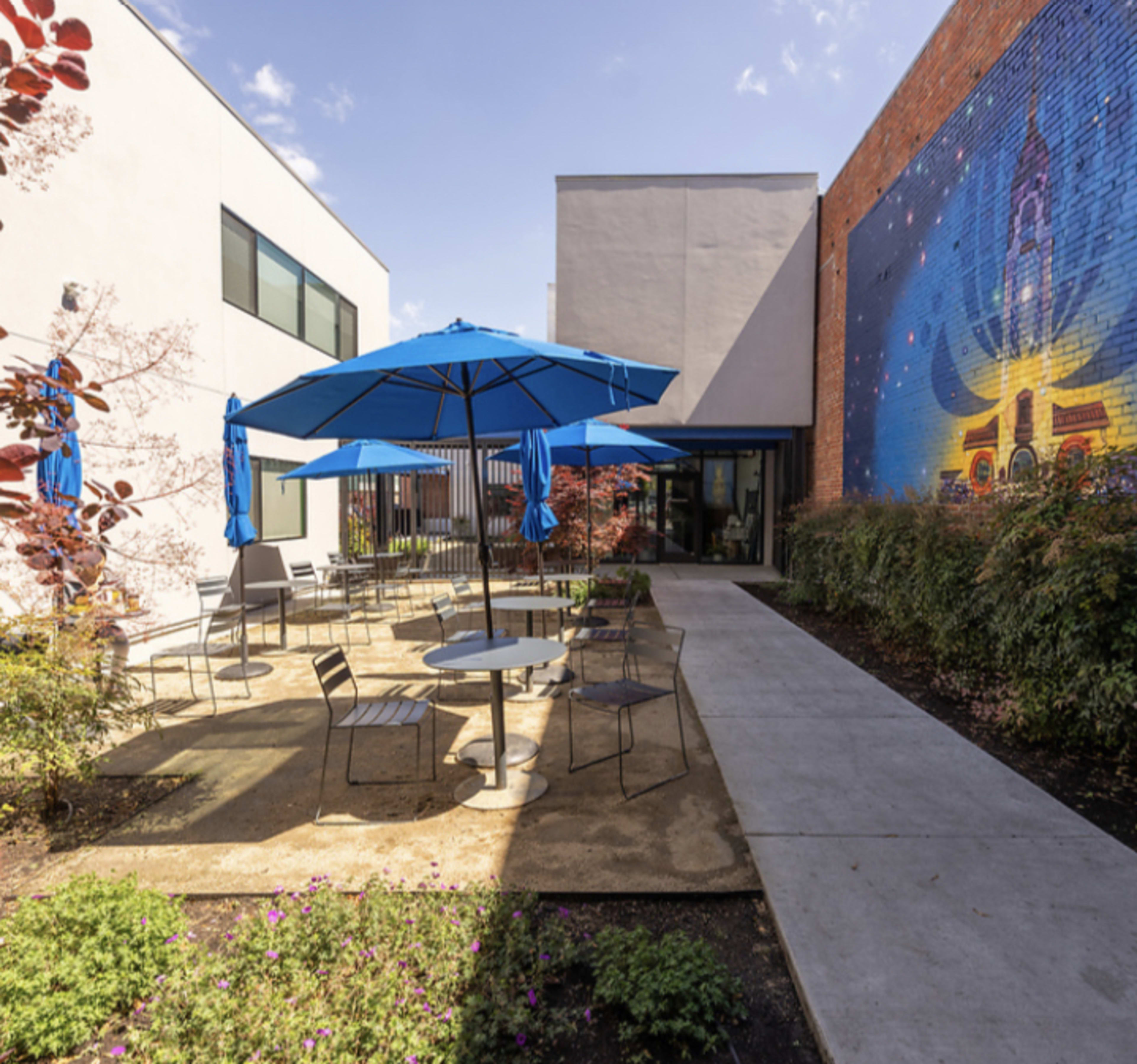 A courtyard with several blue umbrellas over round tables surrounded by greenery and a mural on the wall.