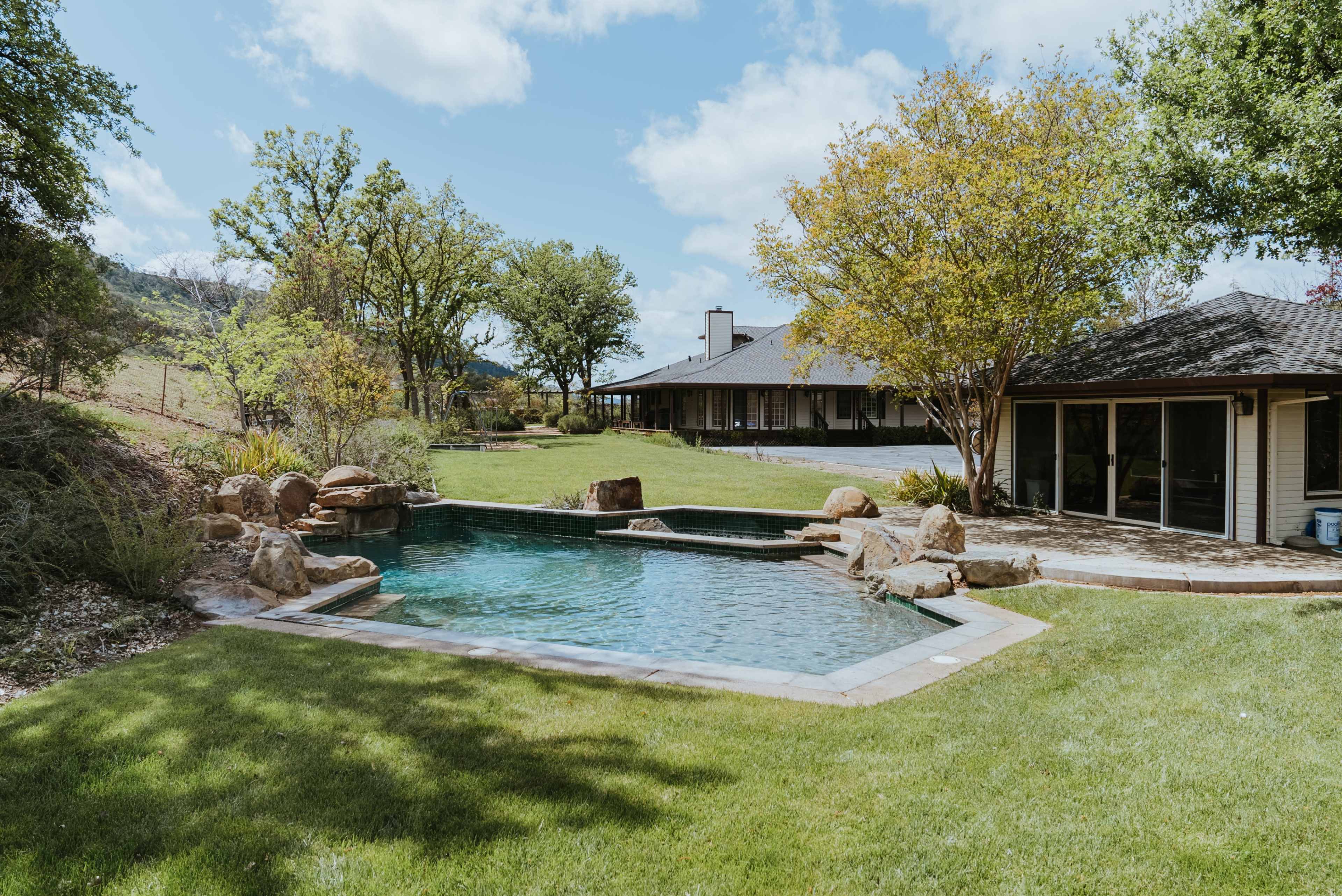 The image shows a backyard with a swimming pool surrounded by greenery and a house in the background.