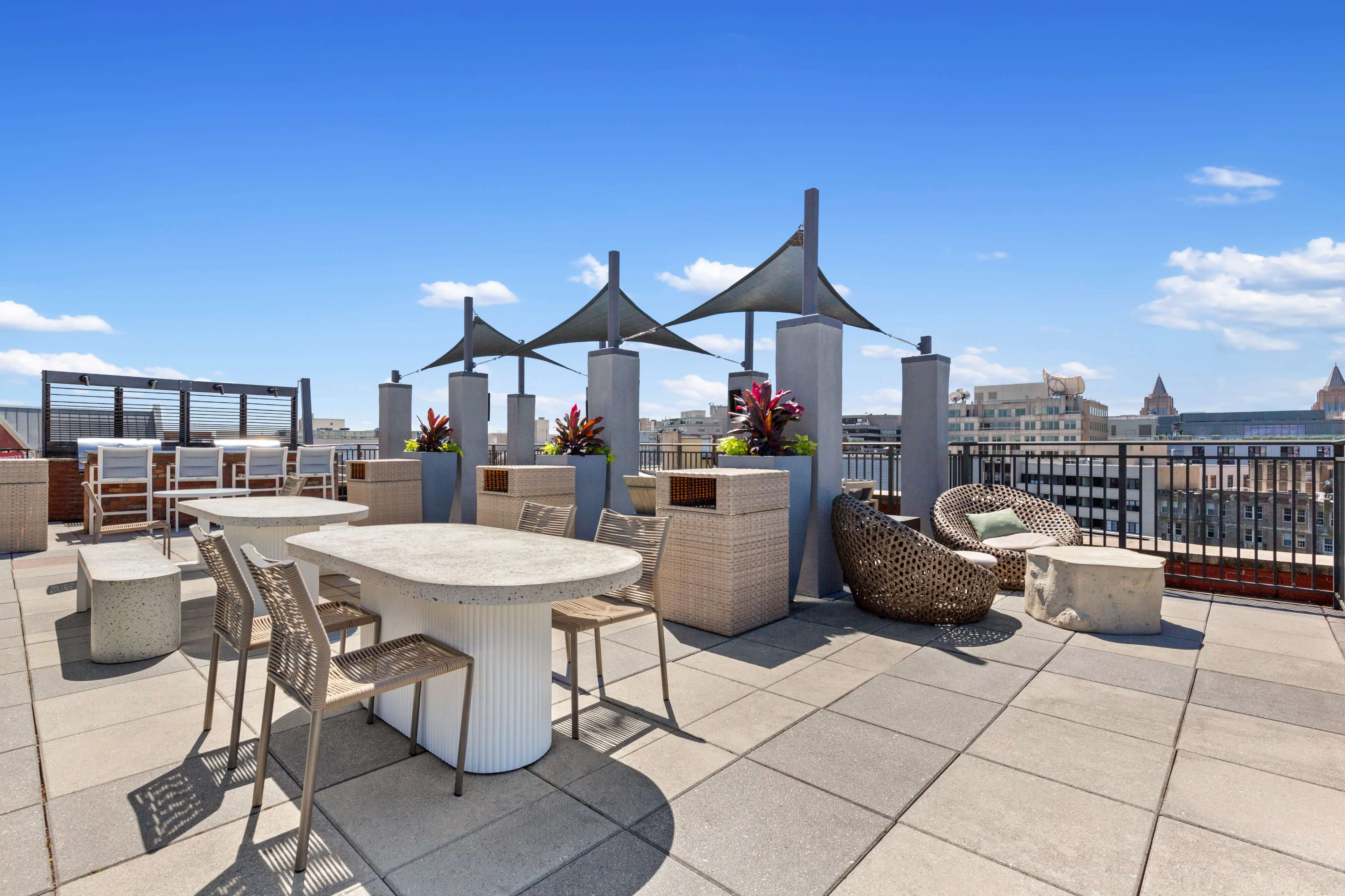 The image shows a rooftop patio featuring various seating arrangements, including a circular table, chairs, and lounge chairs, surrounded by decorative plants and a skyline view.