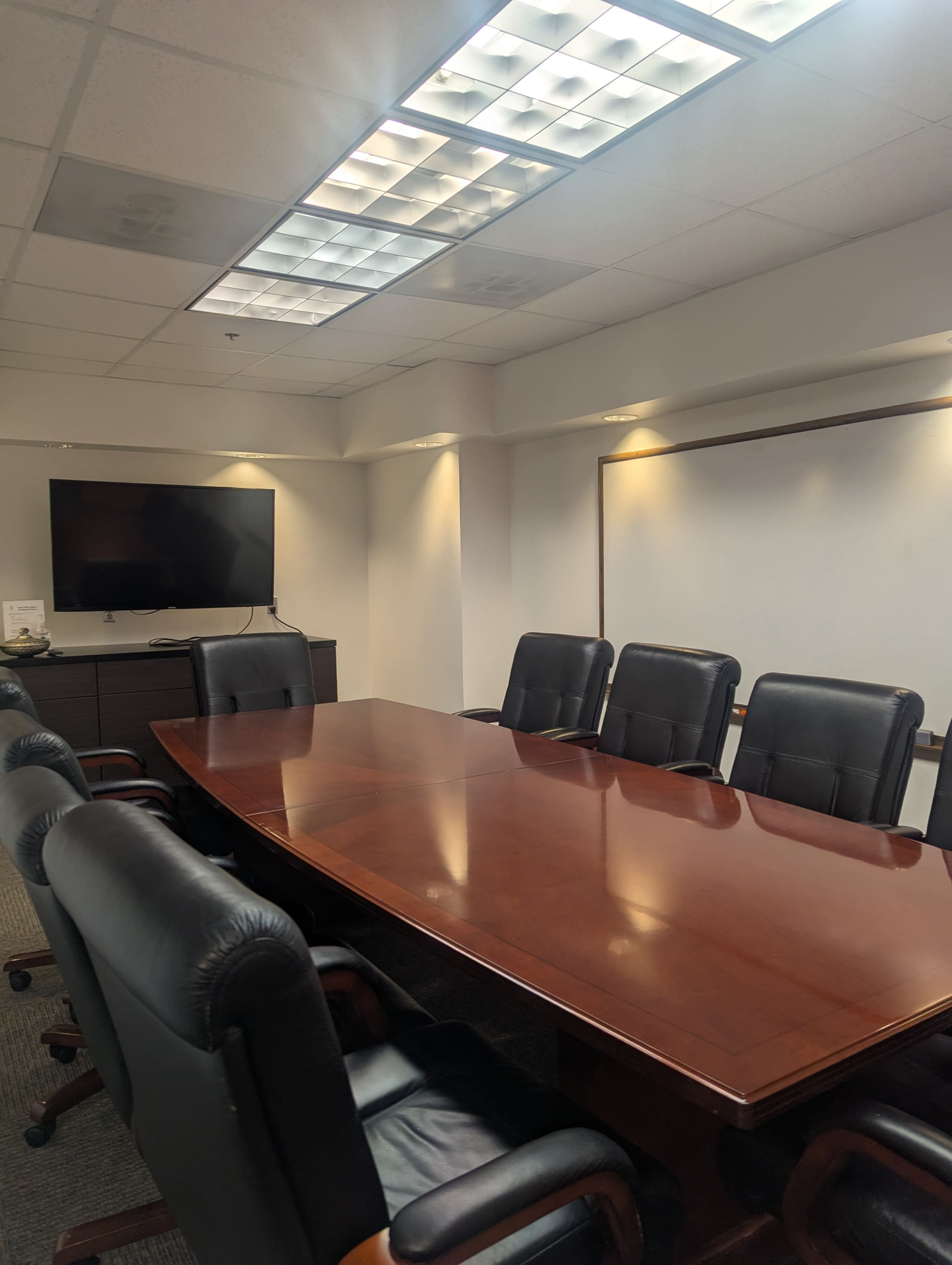 A modern conference room features a long wooden table surrounded by black leather chairs, with a television and whiteboard on the wall.
