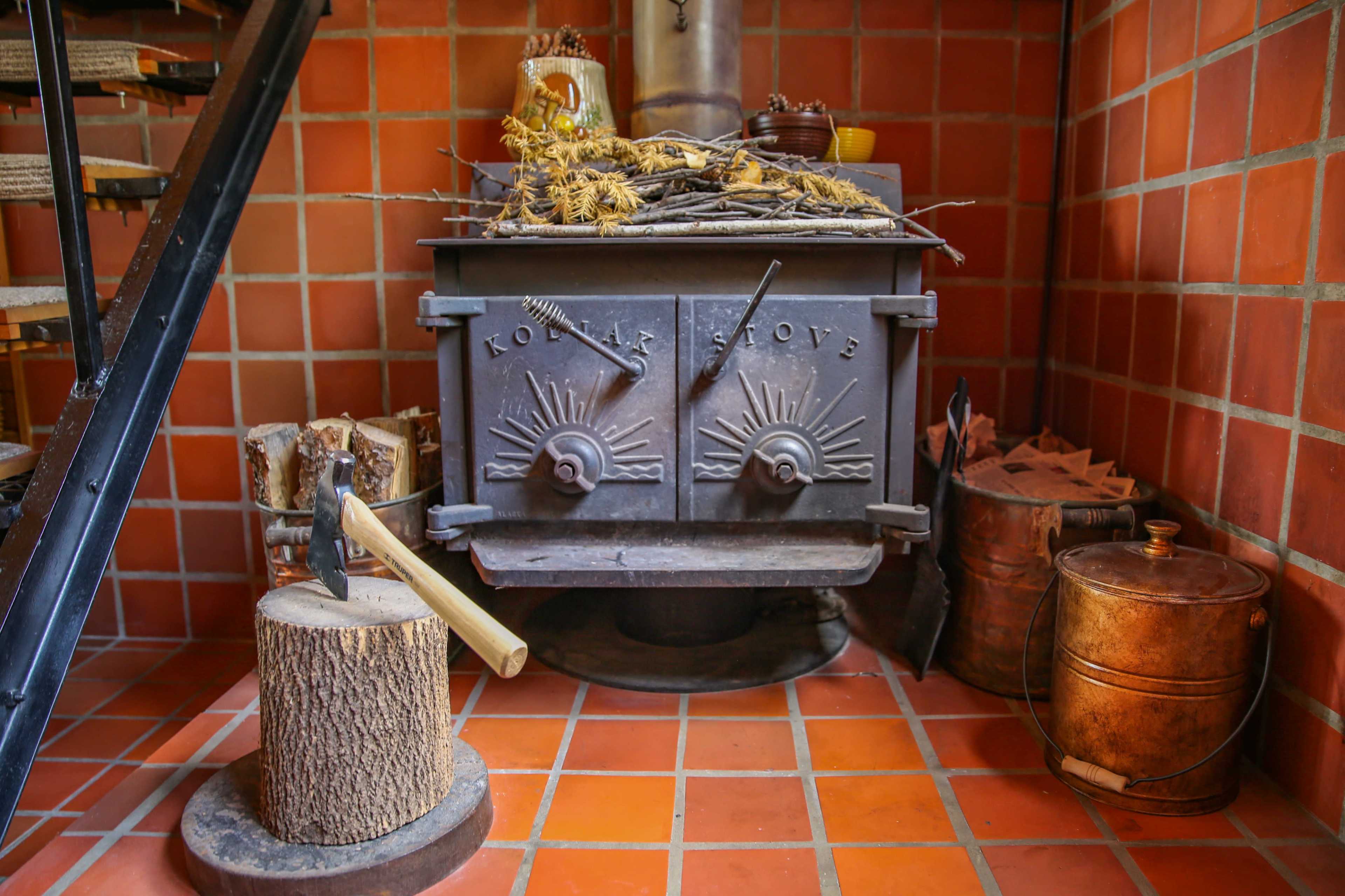 A cast iron wood stove sits in a brick-lined corner, accompanied by a chopping block and a stack of firewood.