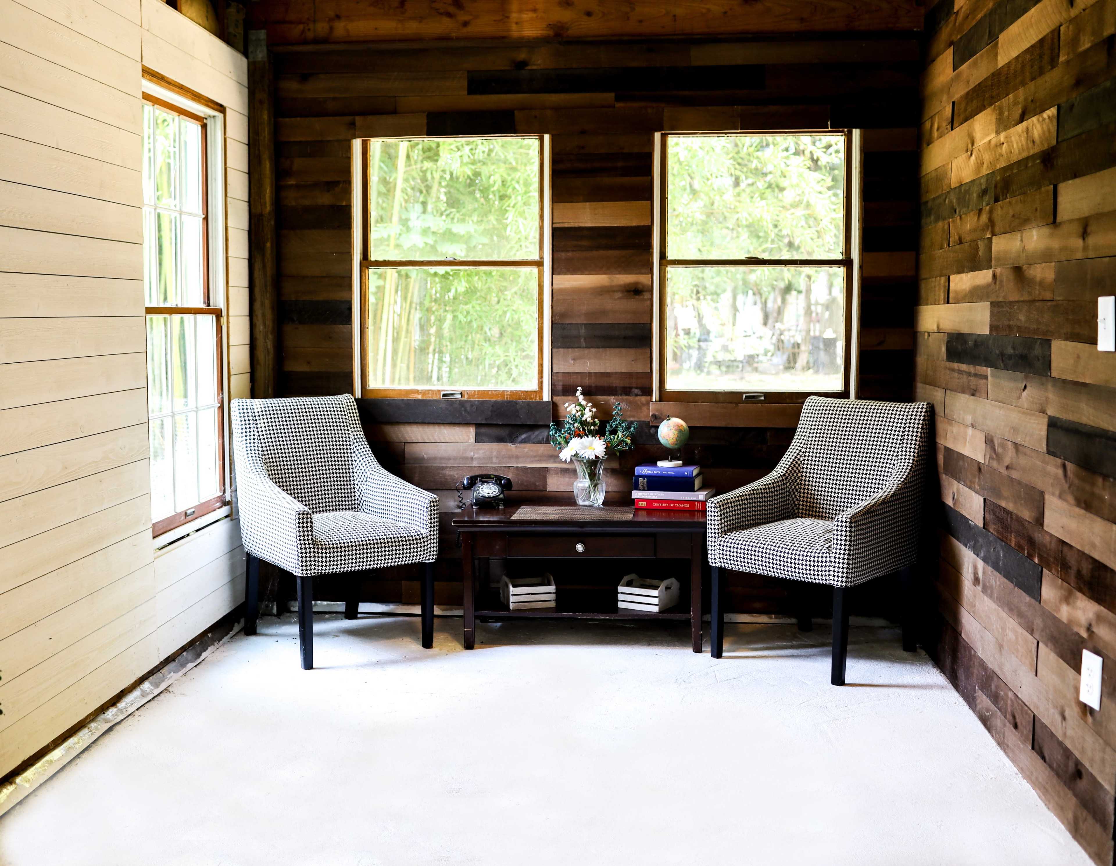A cozy room with two patterned chairs, a dark wooden coffee table, and large windows that provide a view of greenery outside.
