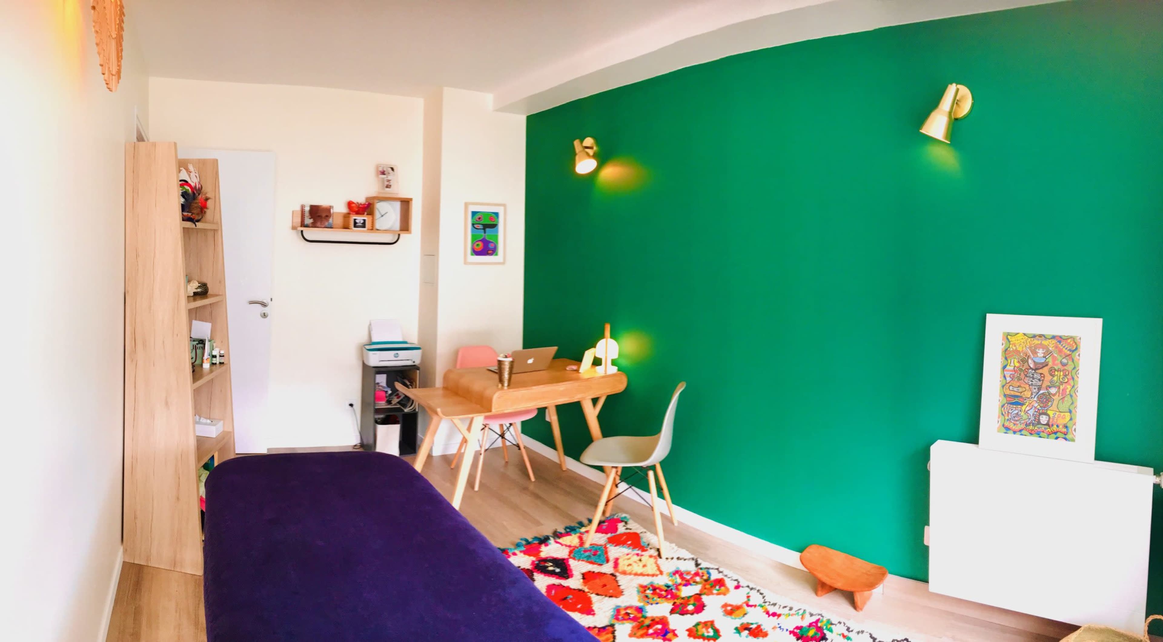 The image shows a small room featuring a green accent wall, a wooden table with a laptop, a bookshelf, and colorful decor on the floor.