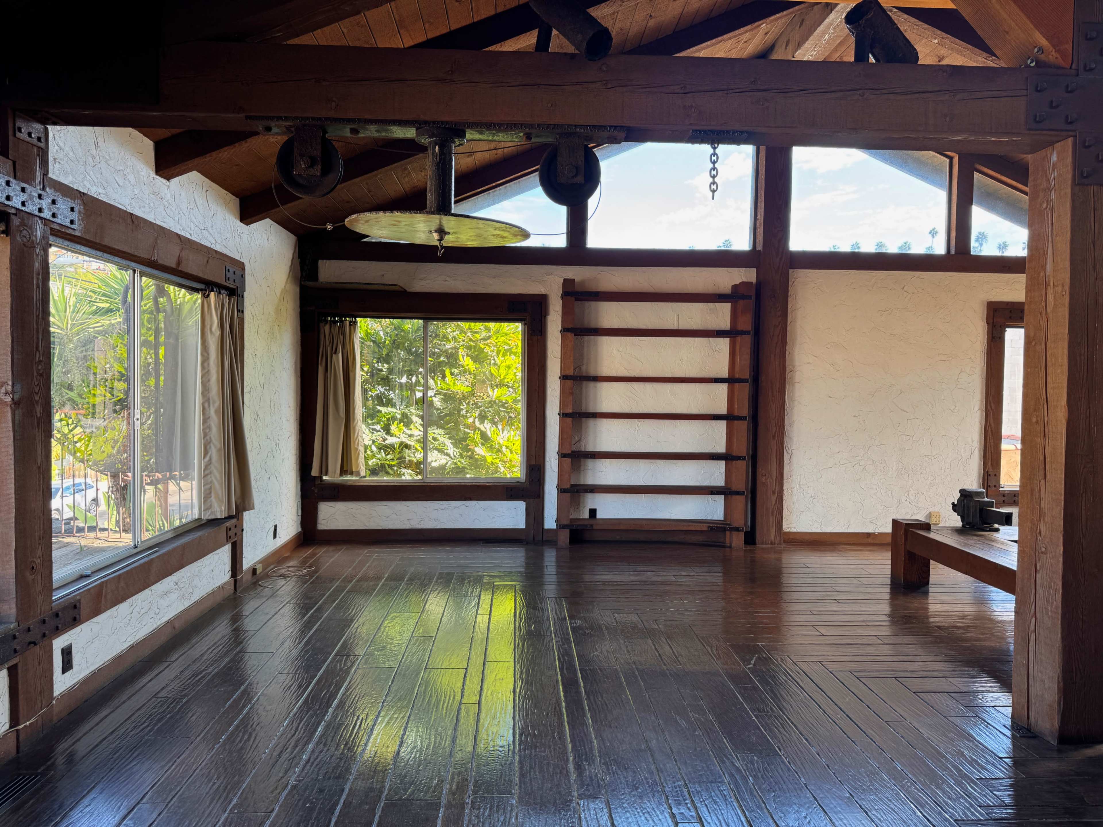 The image shows an interior of a rustic room featuring wooden beams, large windows with views of greenery, and empty shelves against the walls.