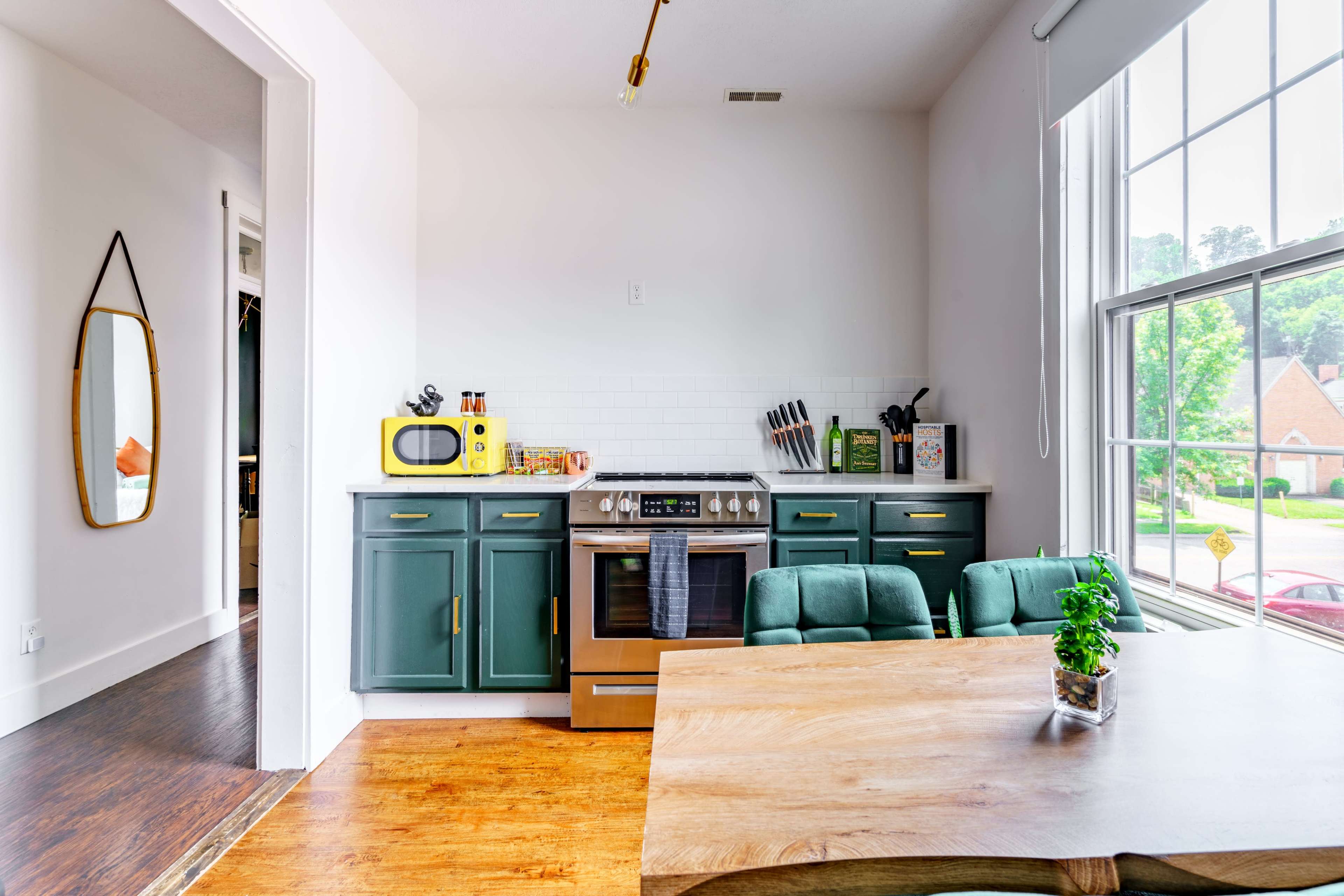 A modern kitchen features green cabinetry, a stainless steel stove, and a wooden dining table, with large windows allowing natural light to illuminate the space.