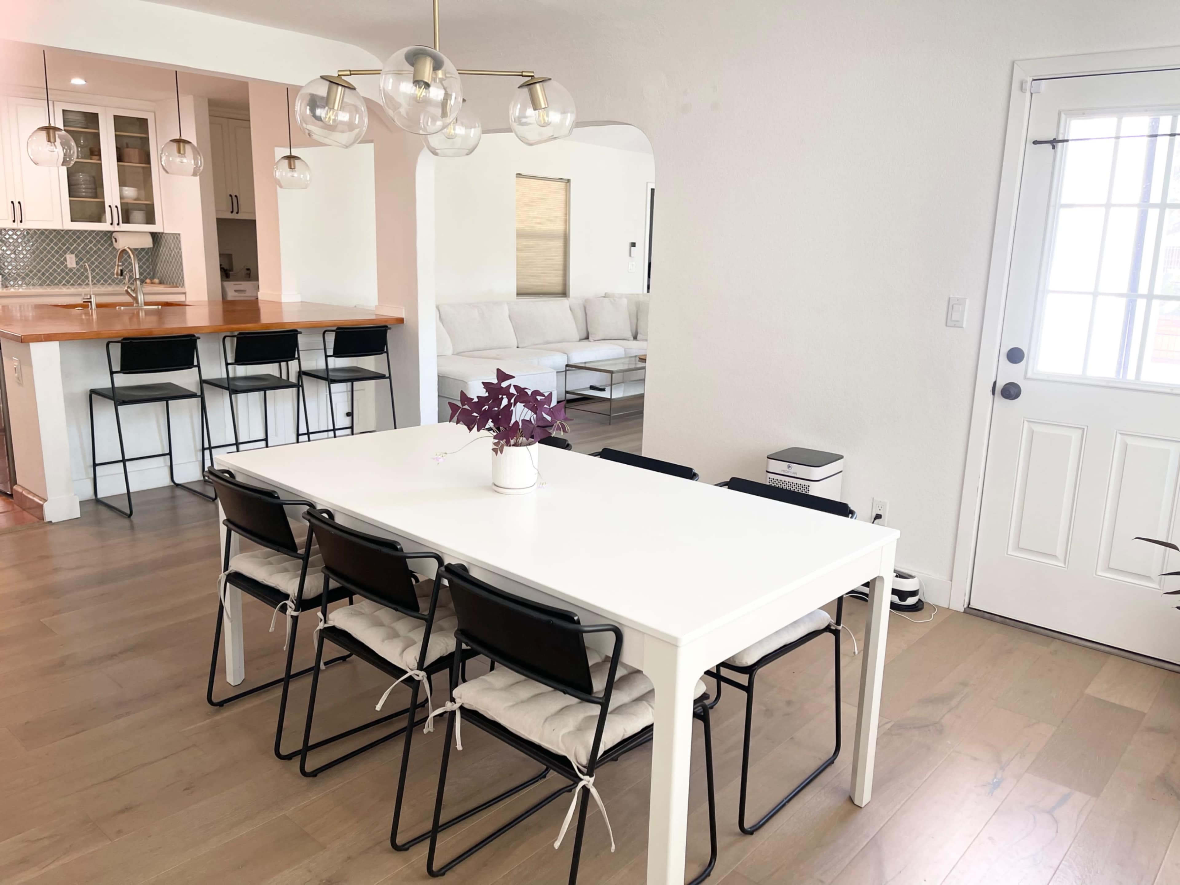 A dining area features a rectangular white table with six black chairs, a small plant centerpiece, and an adjoining kitchen visible in the background.