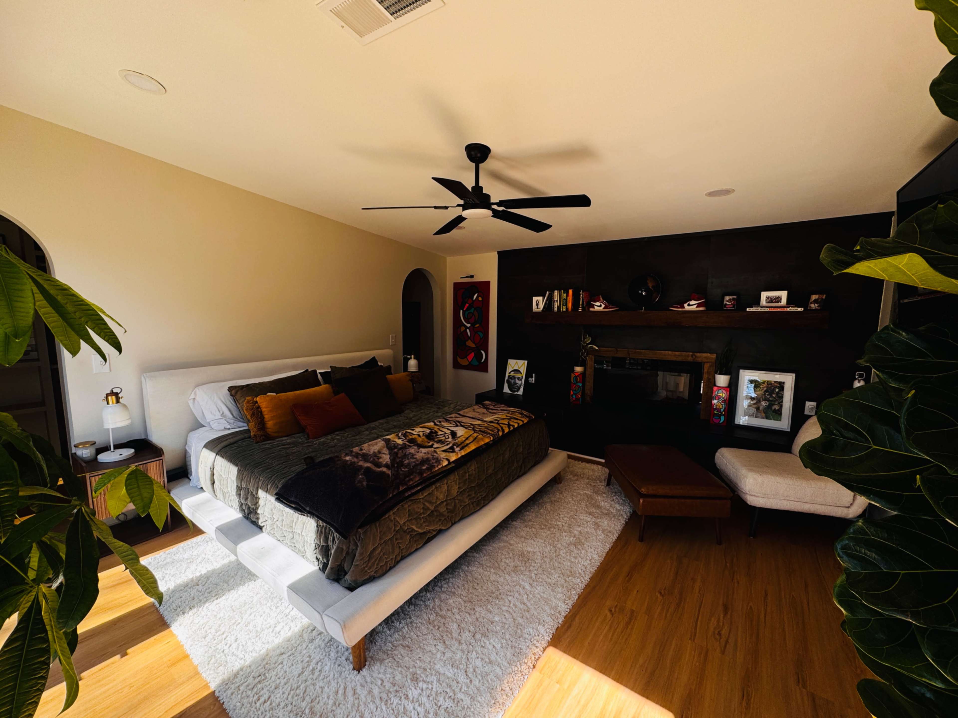 The image shows a well-decorated bedroom featuring a large bed with a dark throw blanket, a fan overhead, a cozy seating area, and a fireplace adorned with various art pieces.