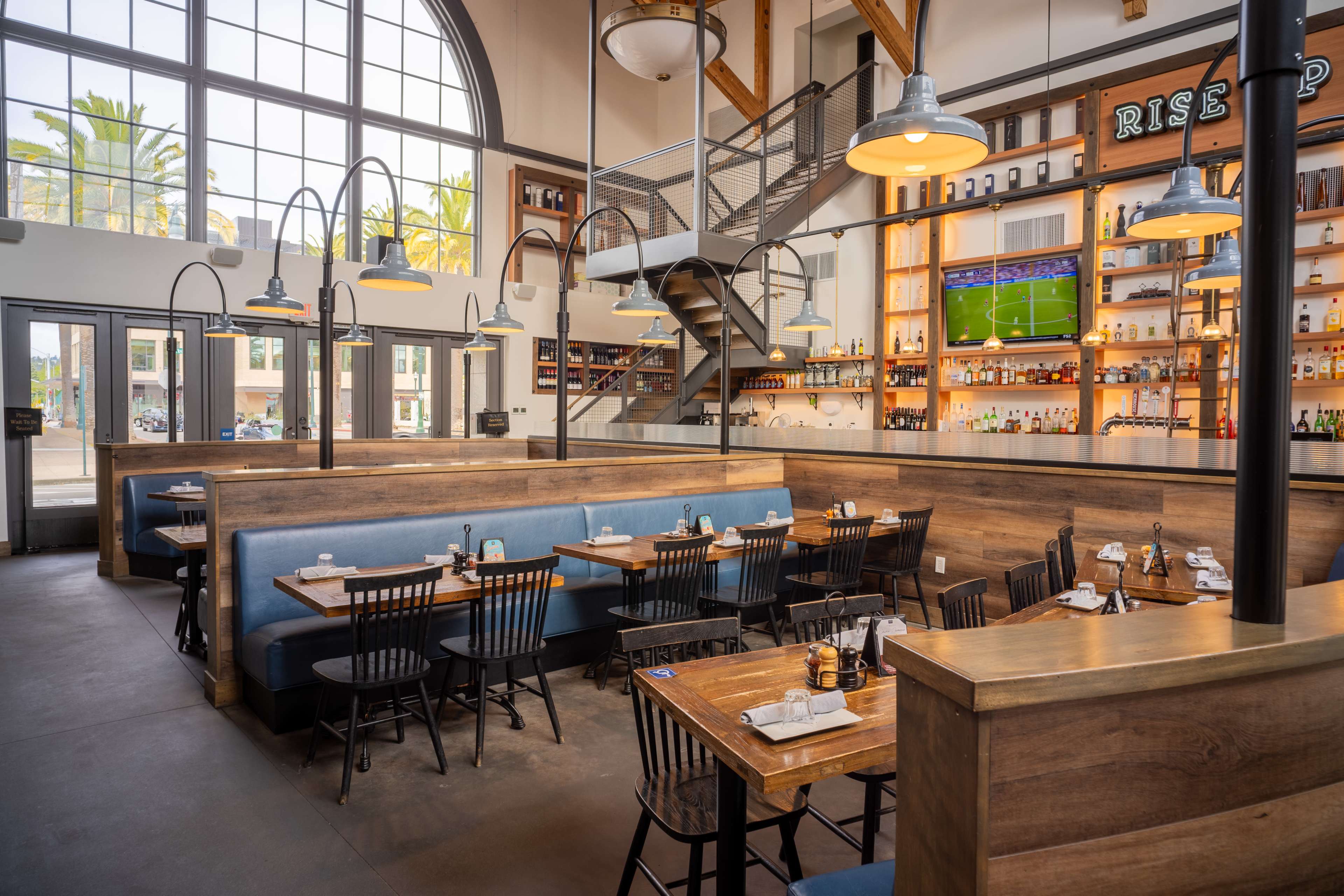 The image shows a modern restaurant interior featuring wooden booths, a bar area with shelves of various bottles, and large windows letting in natural light.