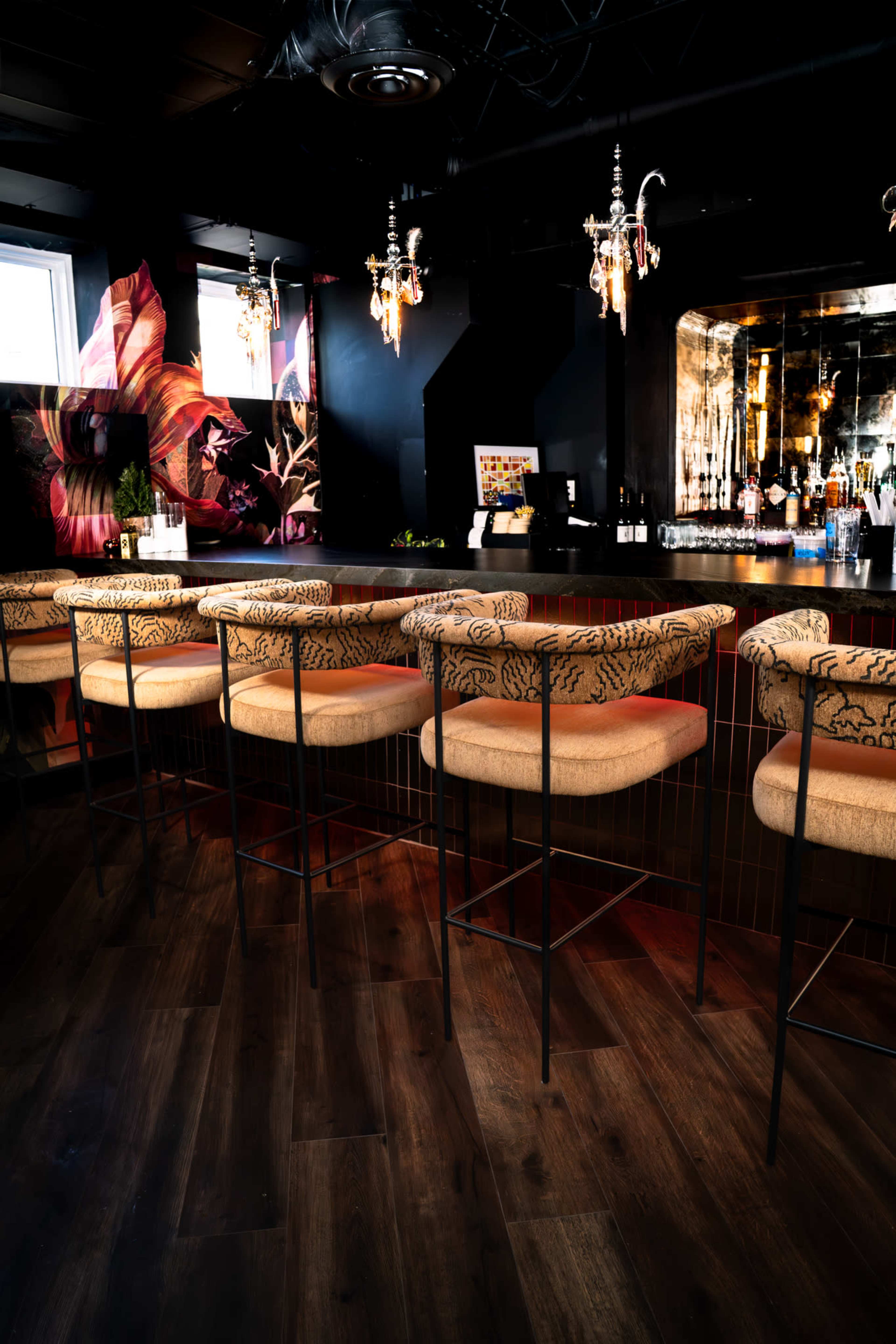 The image shows a chic bar interior with seated stools along a counter, decorative chandeliers, and a vibrant mural on the wall.