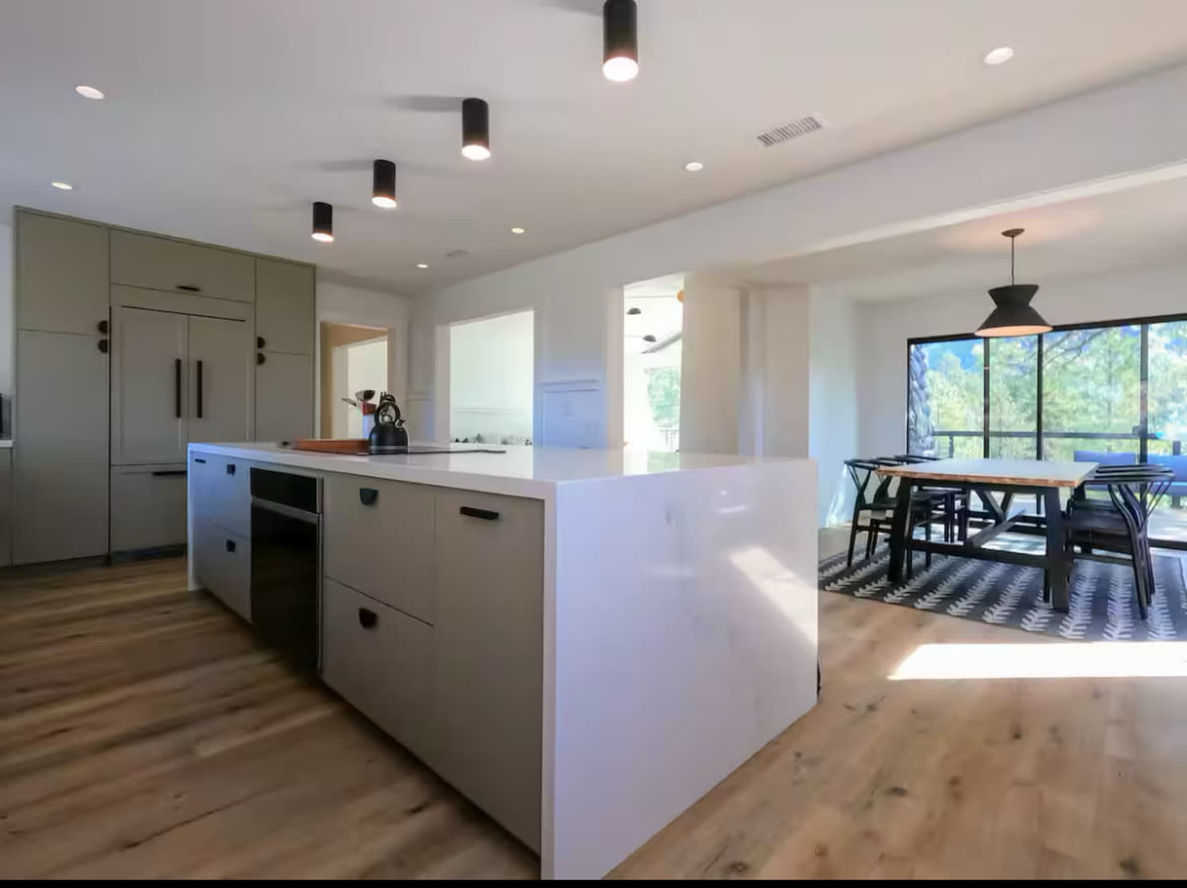 The image shows a modern kitchen featuring a large island with seating, sleek cabinetry, and a dining area with a table and chairs near a window.