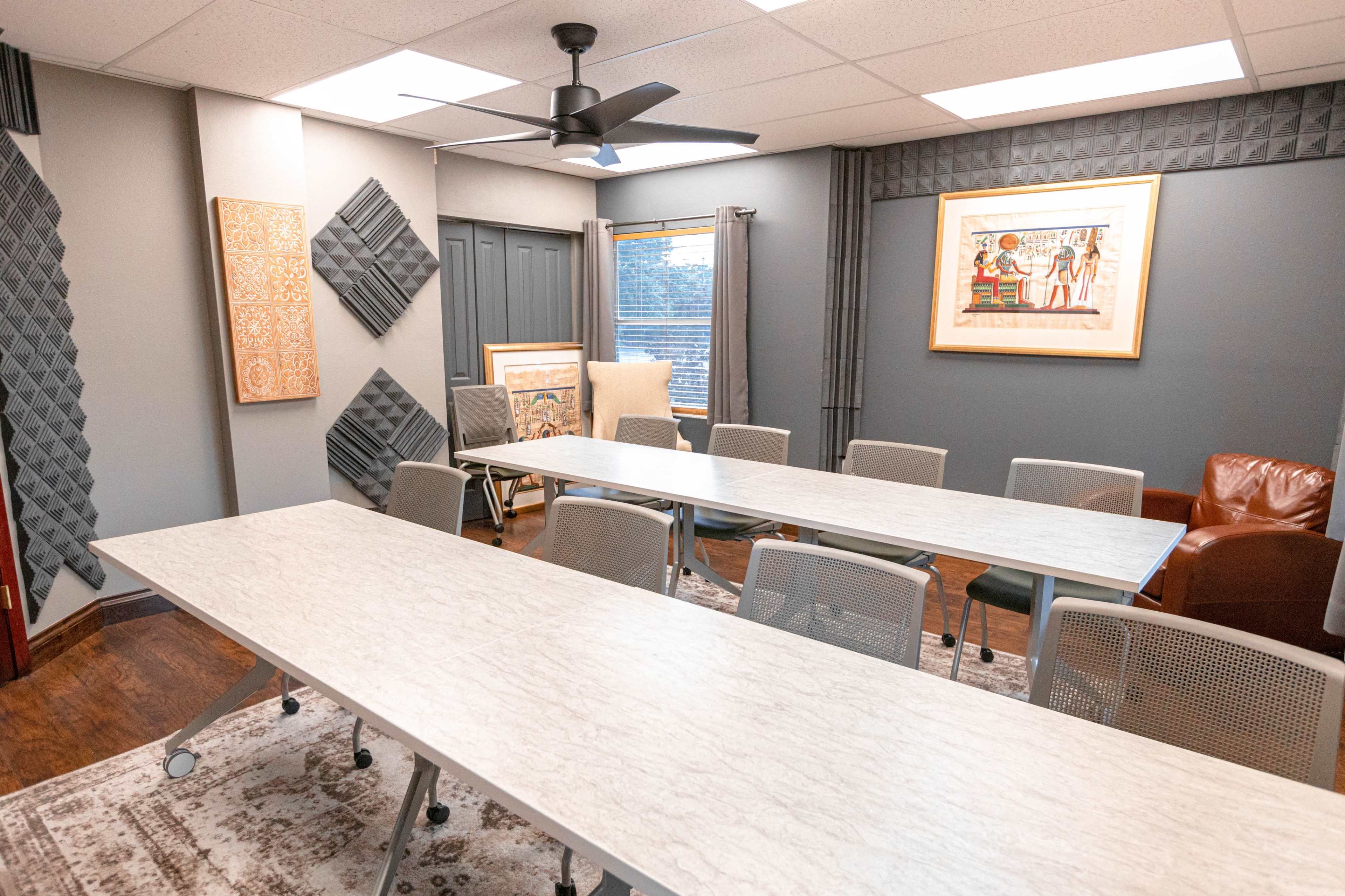 A conference room features multiple long tables arranged in a central layout, surrounded by chairs, with acoustic panels on the walls and artwork displayed.