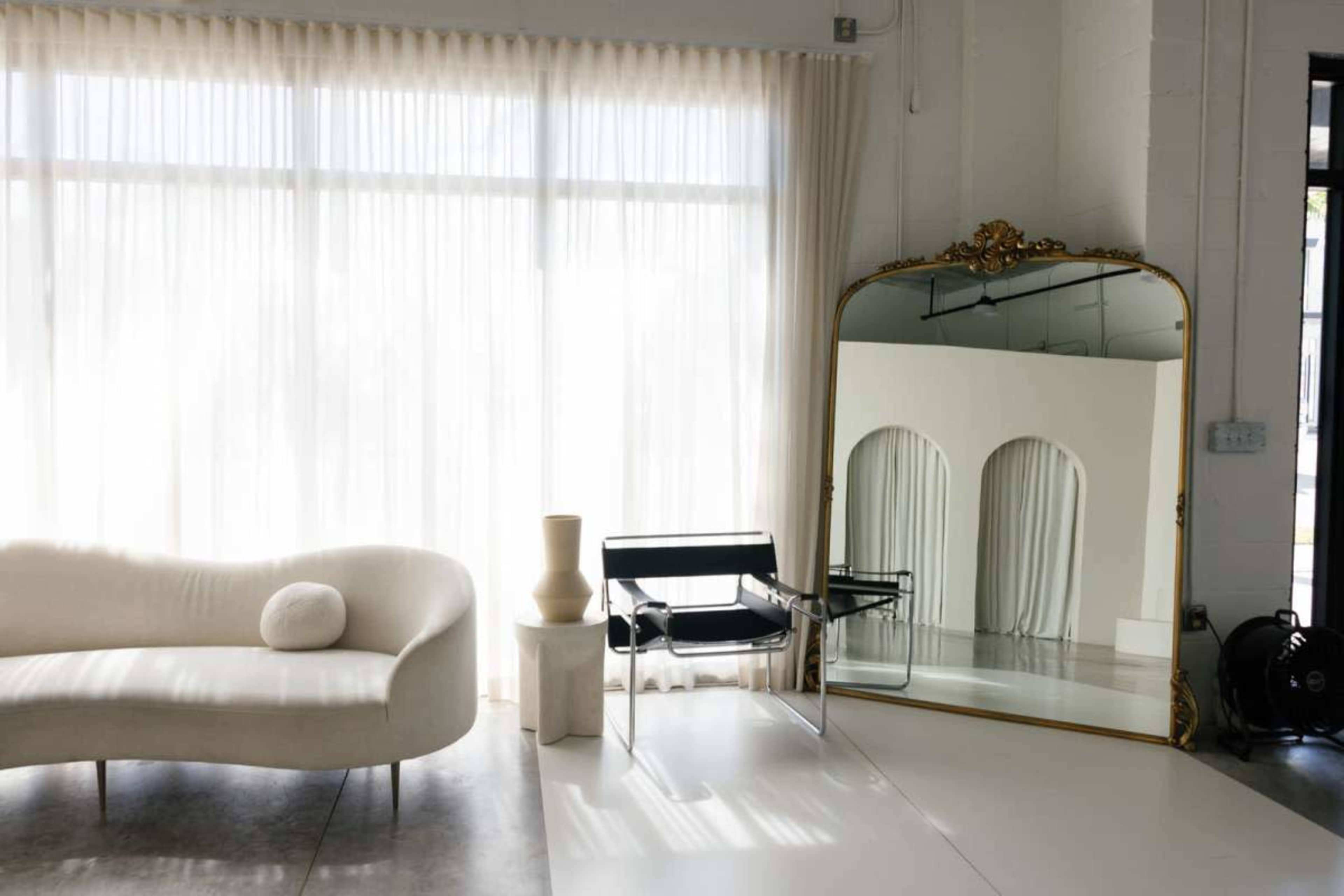 A modern interior features a curved white sofa, a clear acrylic chair, a decorative mirror, and sheer curtains allowing natural light to enter.
