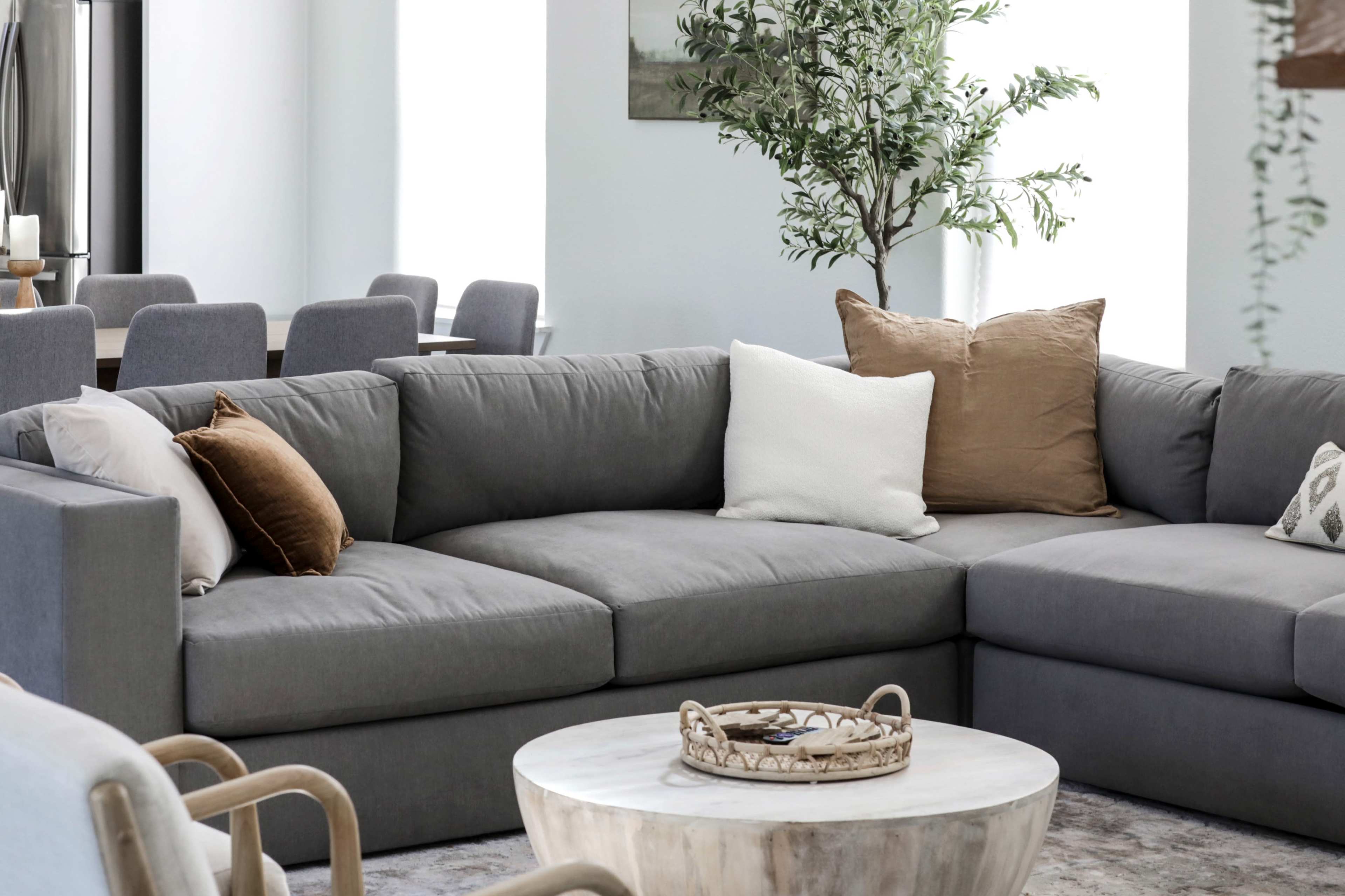 A gray sectional sofa with decorative pillows is positioned next to a round wooden coffee table in a bright living space featuring greenery and dining chairs in the background.