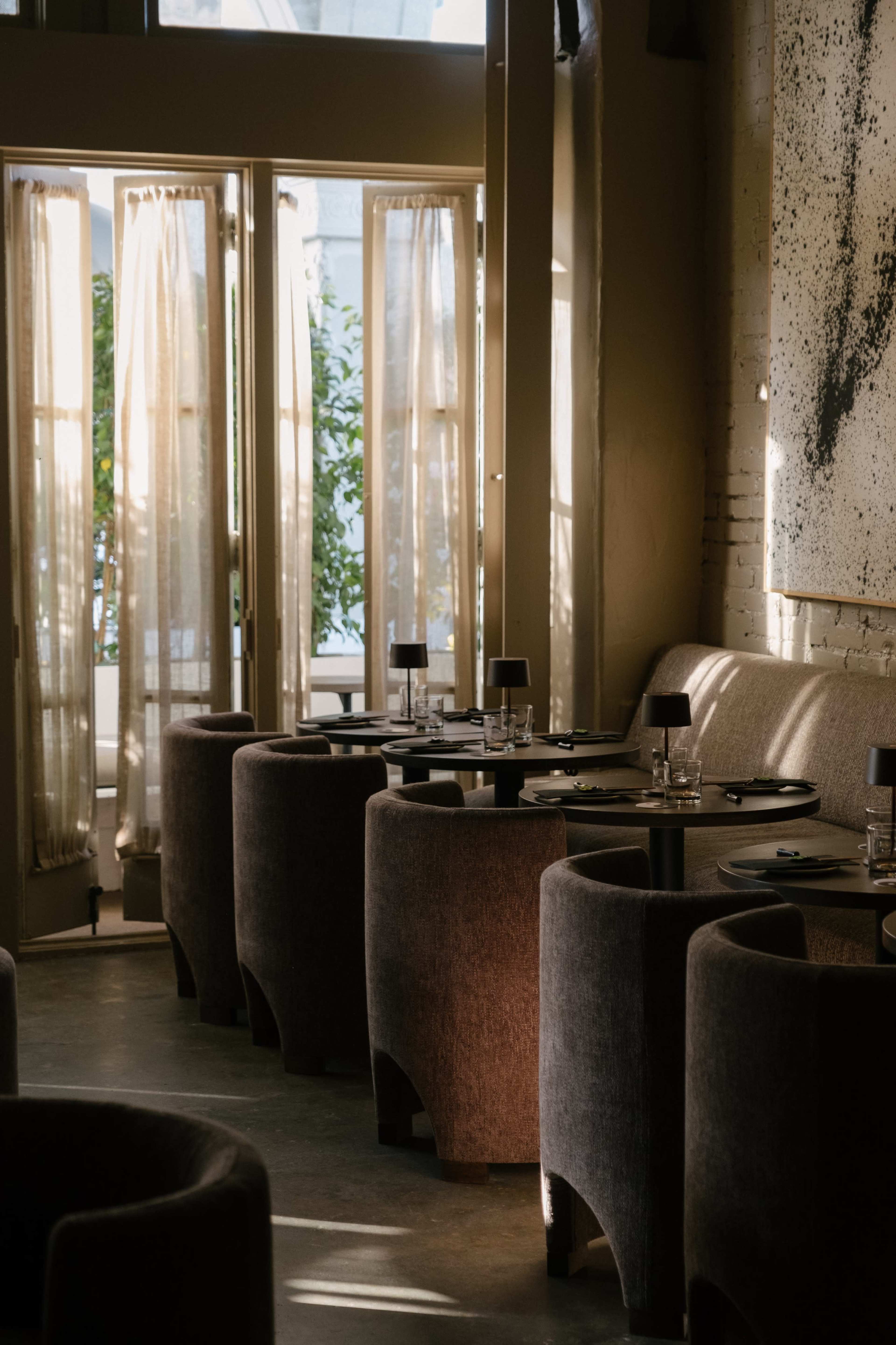 A modern restaurant features round tables with dark plates and glassware, surrounded by plush seating near large, open windows with sheer curtains.