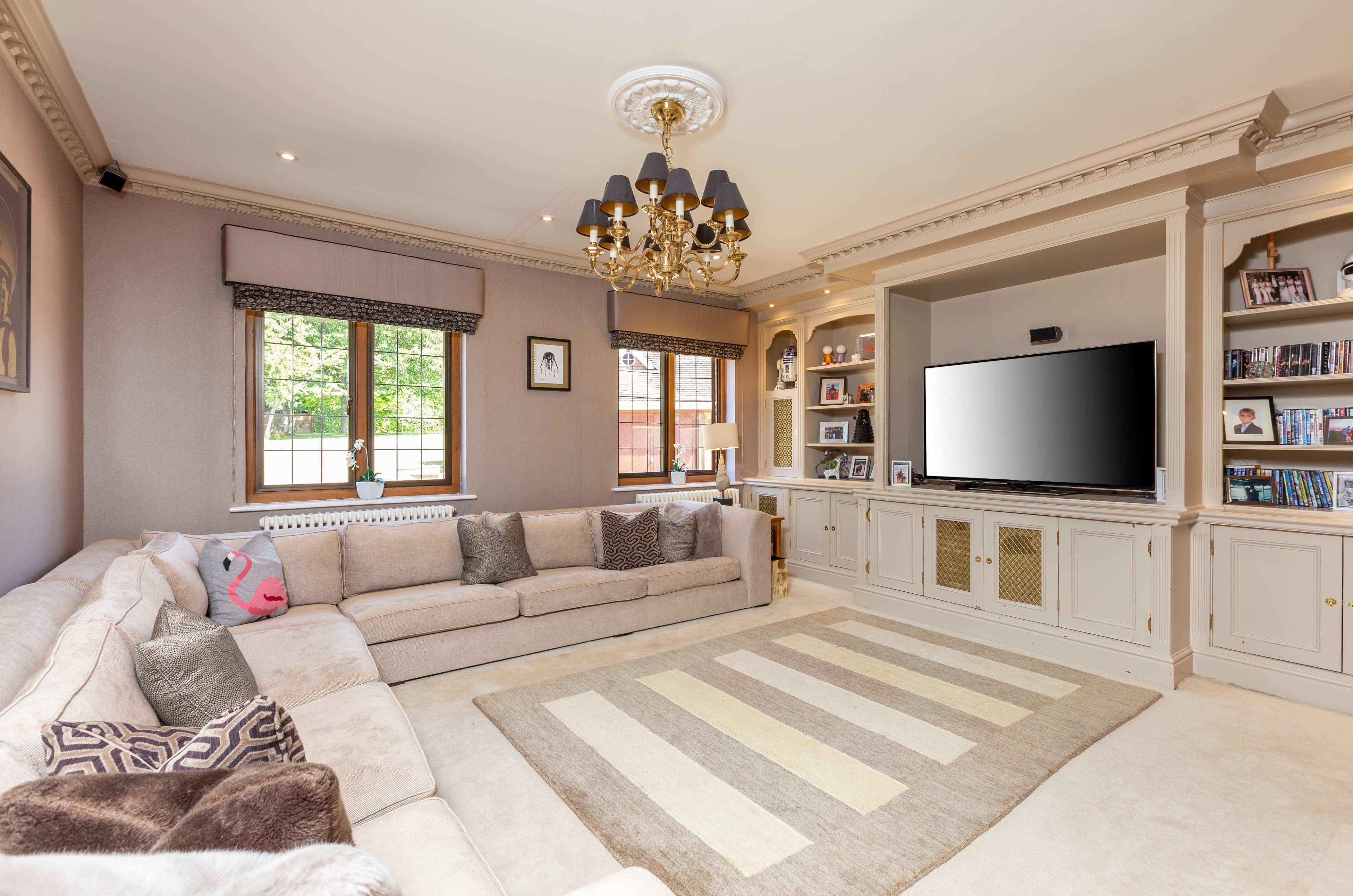 The image shows a spacious living room featuring a large sectional sofa, a patterned area rug, a TV mounted on a light-colored entertainment unit, and decorative shelves filled with books and framed photos.