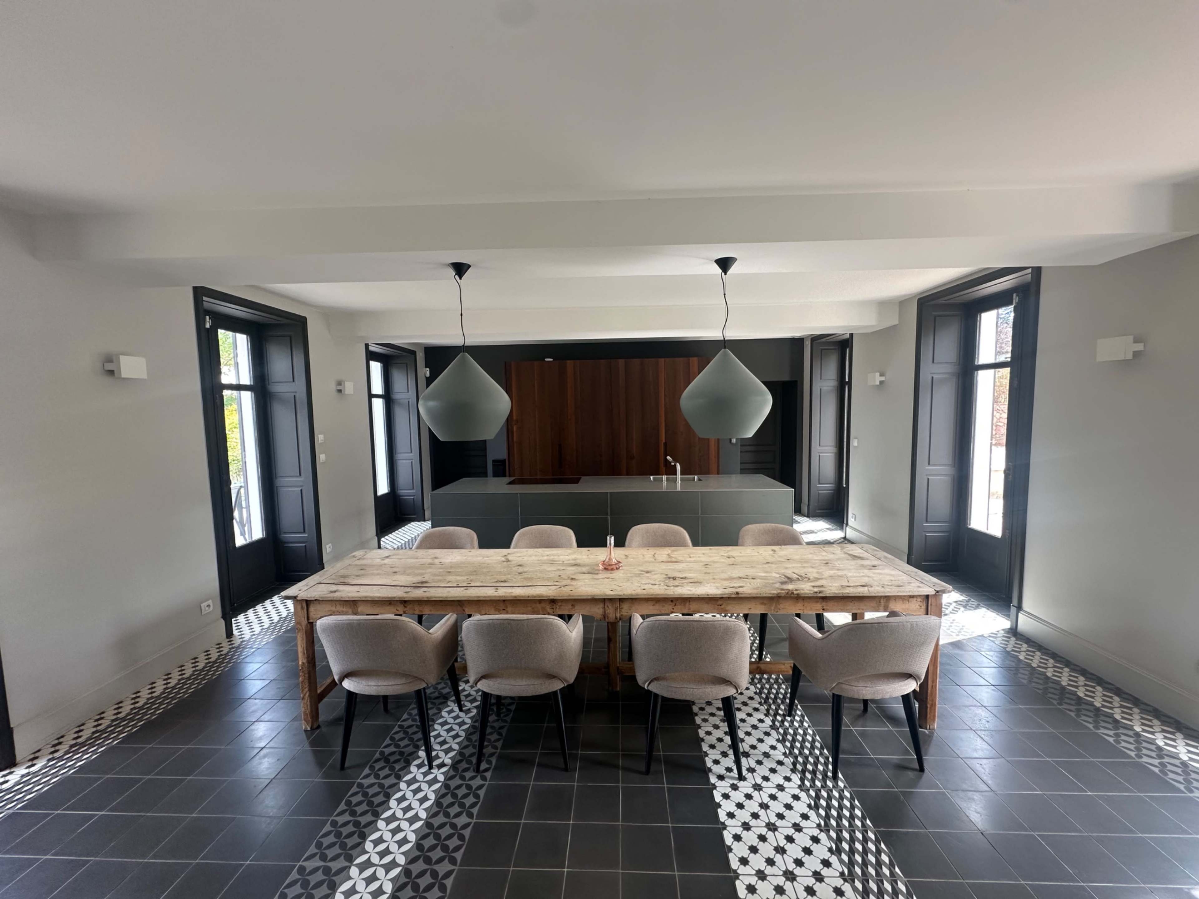 A large wooden dining table with eight chairs is set in a modern room featuring gray walls, pendant lighting, and tiled flooring with a geometric pattern.