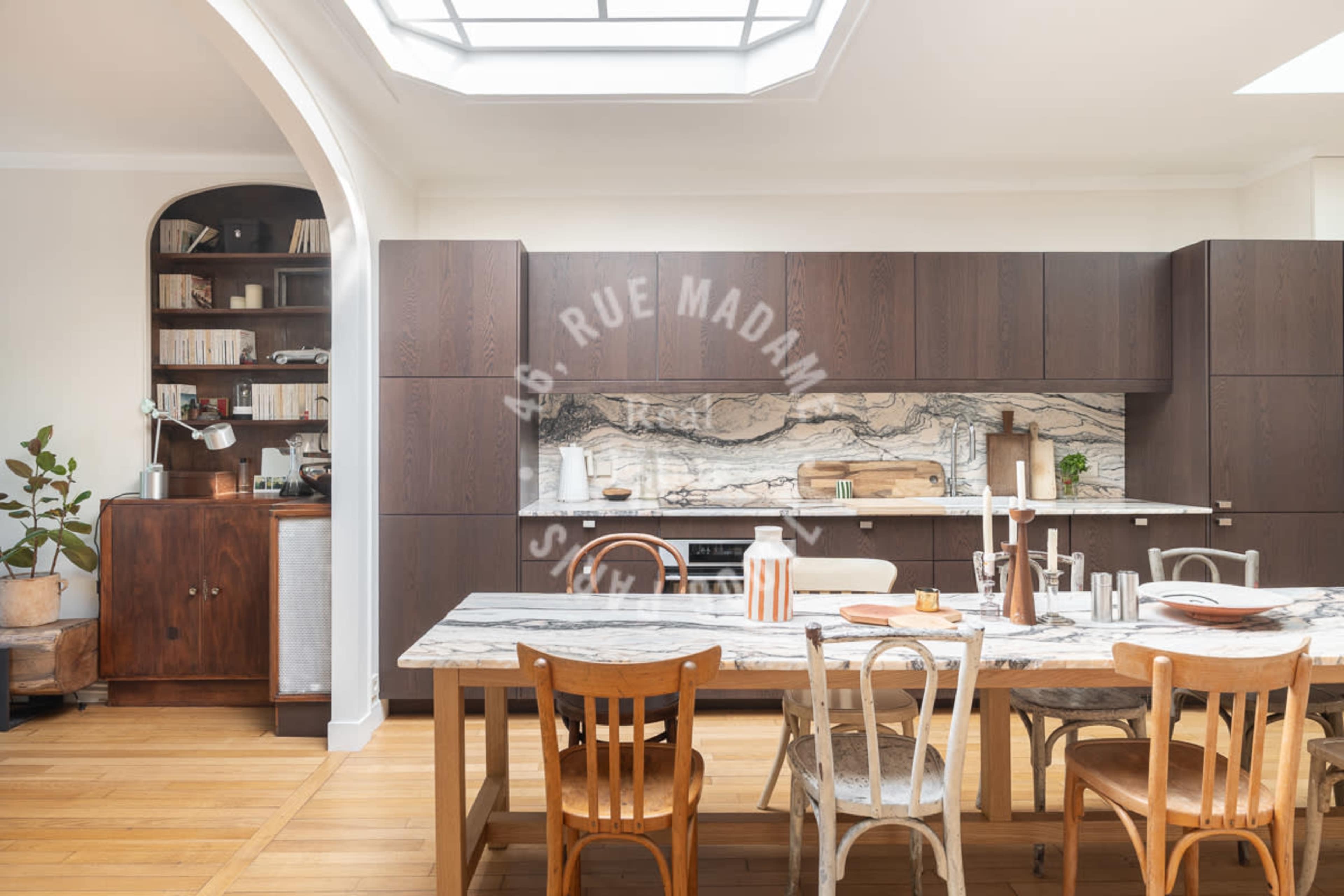 The image features a modern kitchen with a marble countertop, dark wood cabinetry, and a dining table set with plates and utensils, alongside a bookshelf and a potted plant in the background.