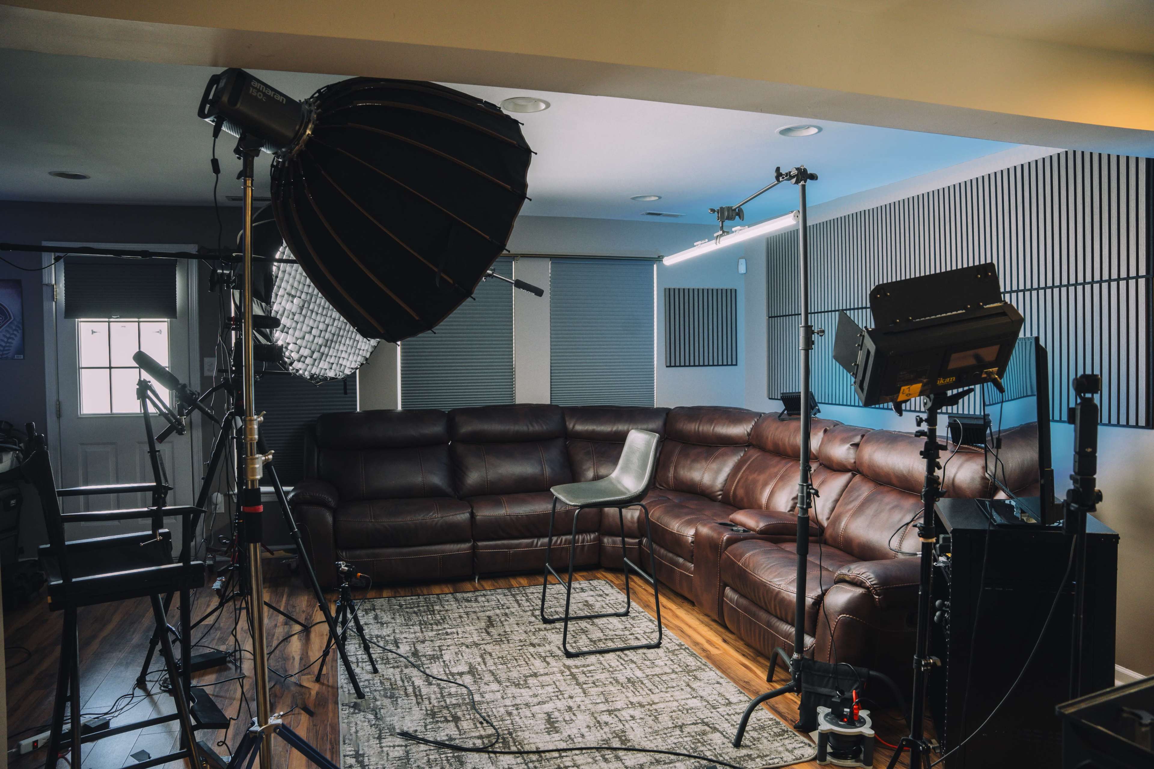 The image shows a well-lit photography studio featuring a brown leather sectional couch, a single empty chair, and various lighting equipment arranged around the space.